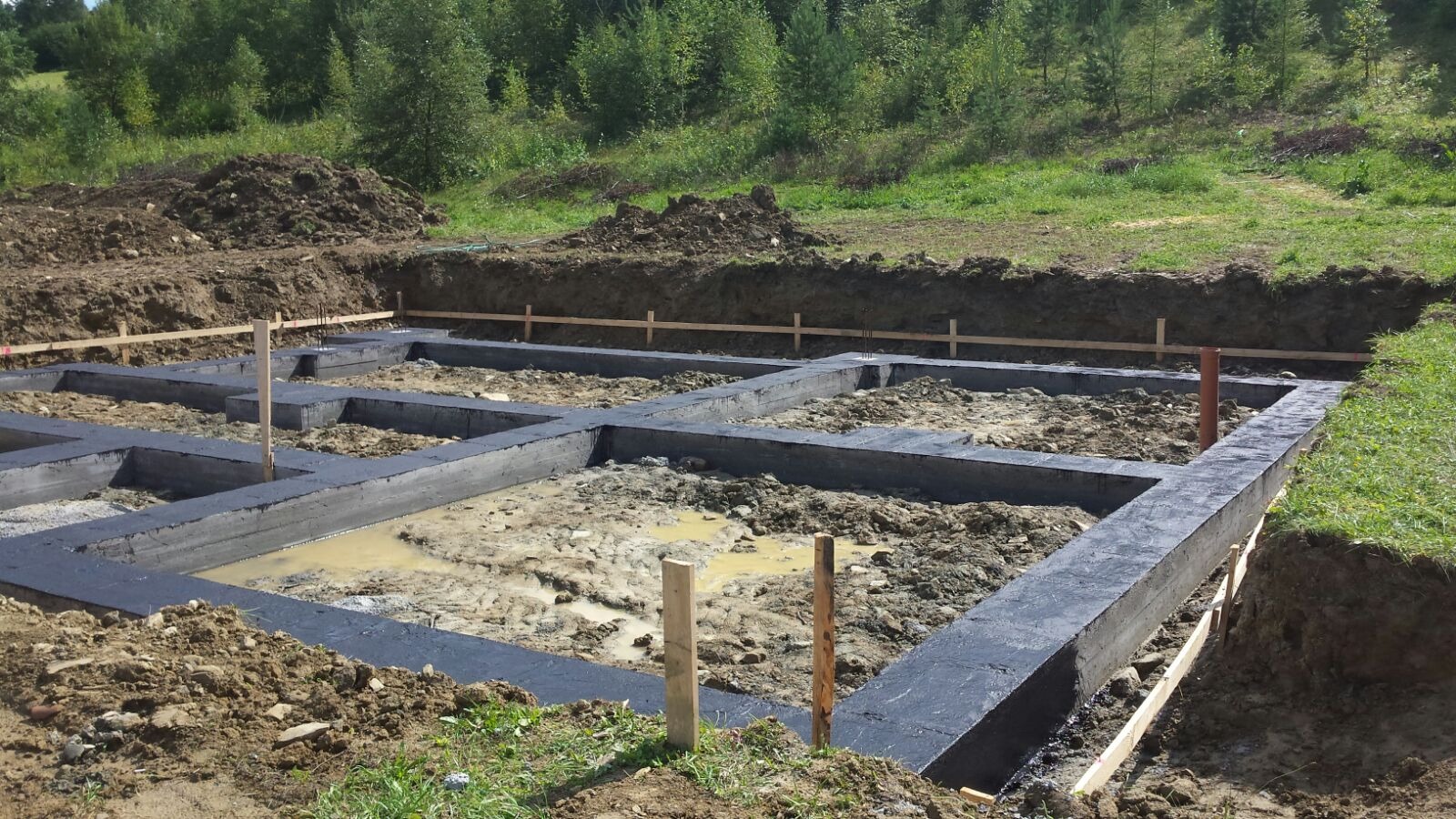 Zalane fundamenty domu jednorodzinnego z hydroizolacją, widoczne drewniane szalunki i wykop ziemny. W tle zieleń drzew i trawy, słoneczny dzień.