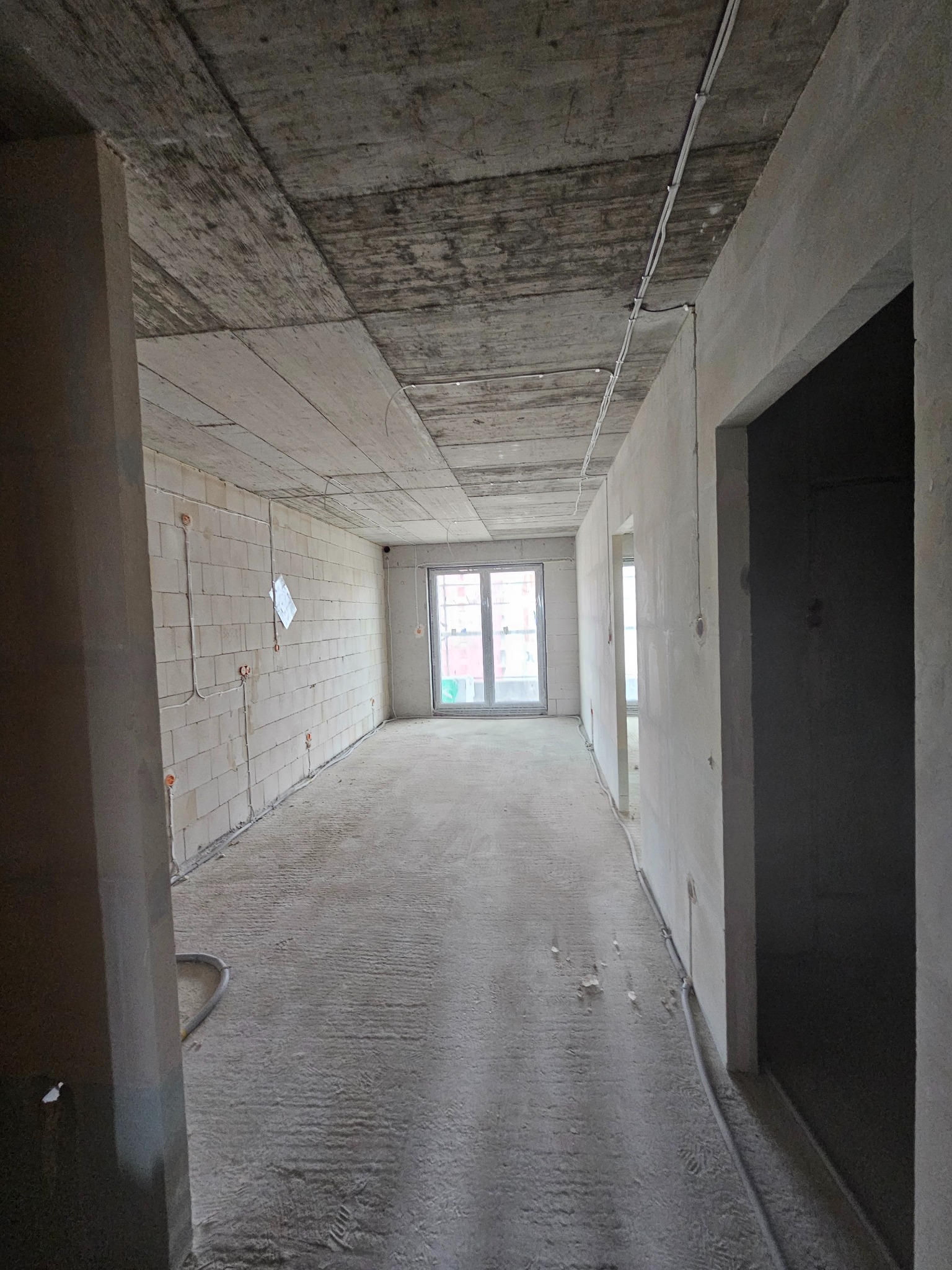 Surowy stan mieszkania w trakcie remontu: widoczne przewody elektryczne na ścianach z pustaków, betonowa podłoga i sufit. Okno w tle.