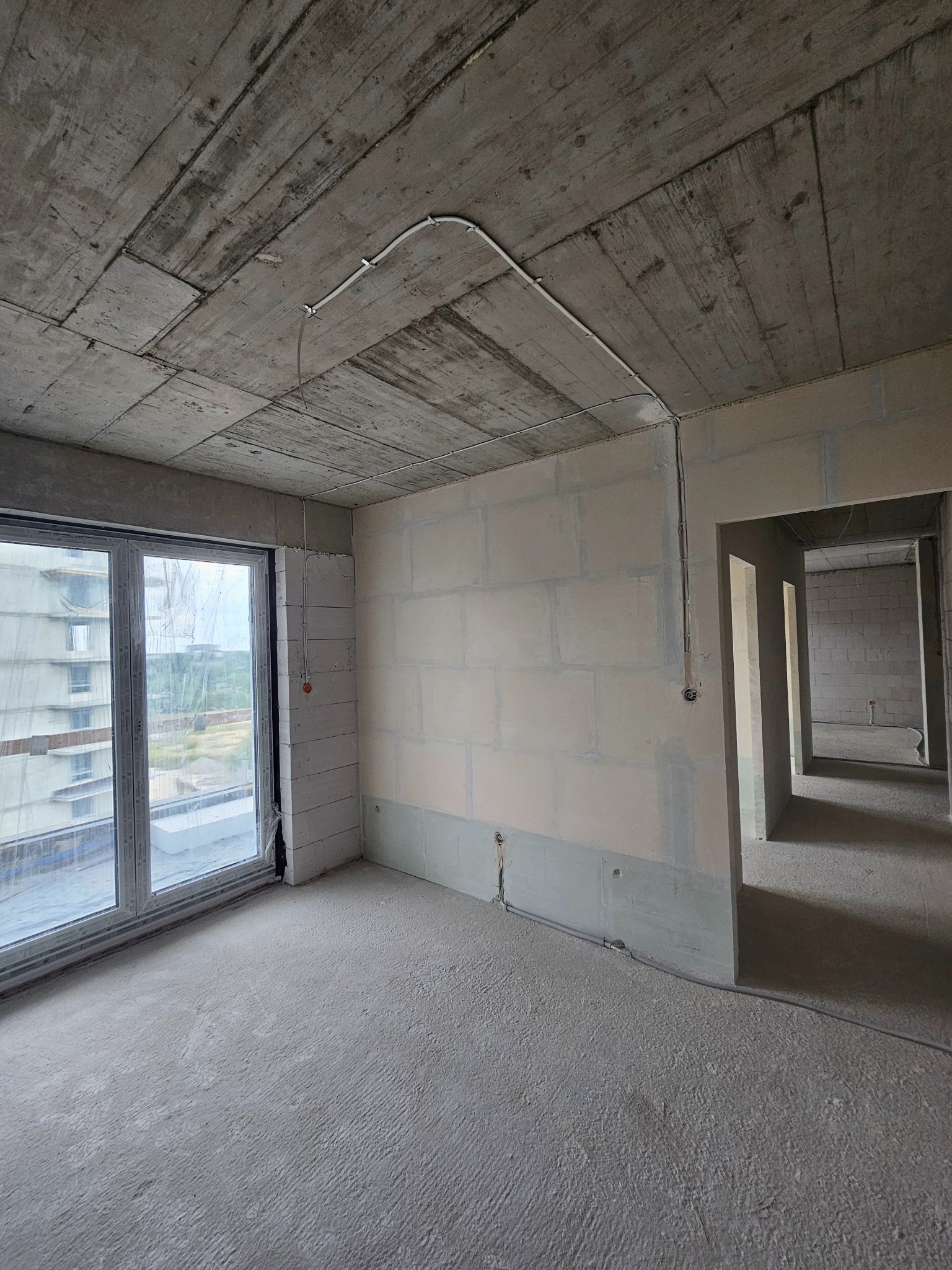 Surowy stan wnętrza: betonowy strop z instalacją, ściany z bloczków, okno z widokiem na budynek. W tle widoczne kolejne pomieszczenia w stanie deweloperskim.