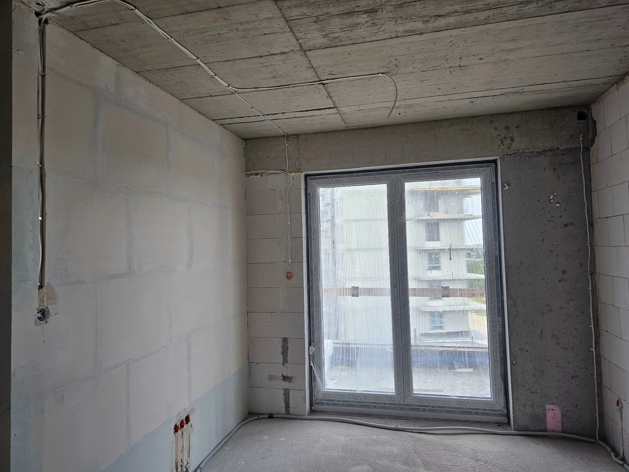 Surowy pokój w trakcie remontu: widoczne przewody elektryczne na ścianach i suficie, okno z widokiem na budowę. Ściany z betonu komórkowego i surowy beton.