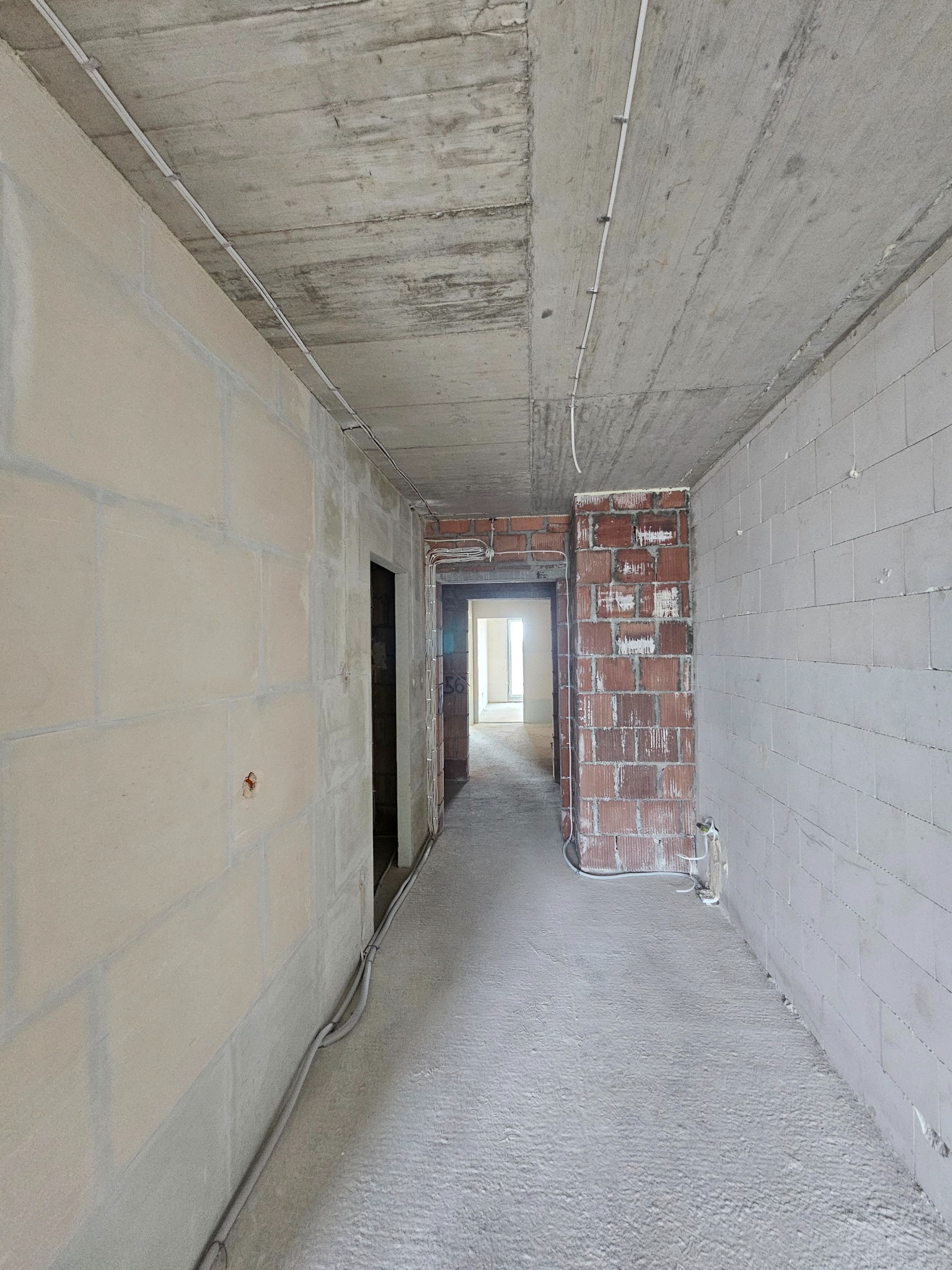 Wnętrze w trakcie remontu: surowy beton sufitu, ściany z bloków i cegieł, widoczne przewody elektryczne. Stan deweloperski z elementami instalacji.