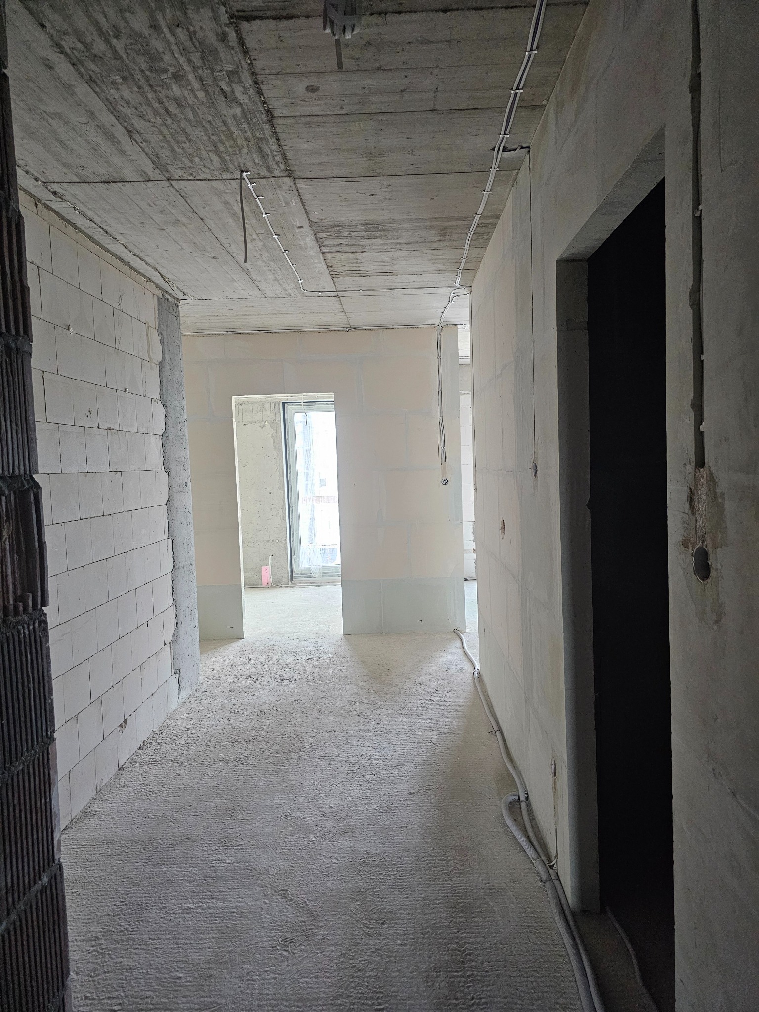 Surowy stan mieszkania w trakcie remontu: widoczne ściany z bloczków, betonowy sufit z przewodami elektrycznymi, w tle okno z widokiem na zewnątrz.