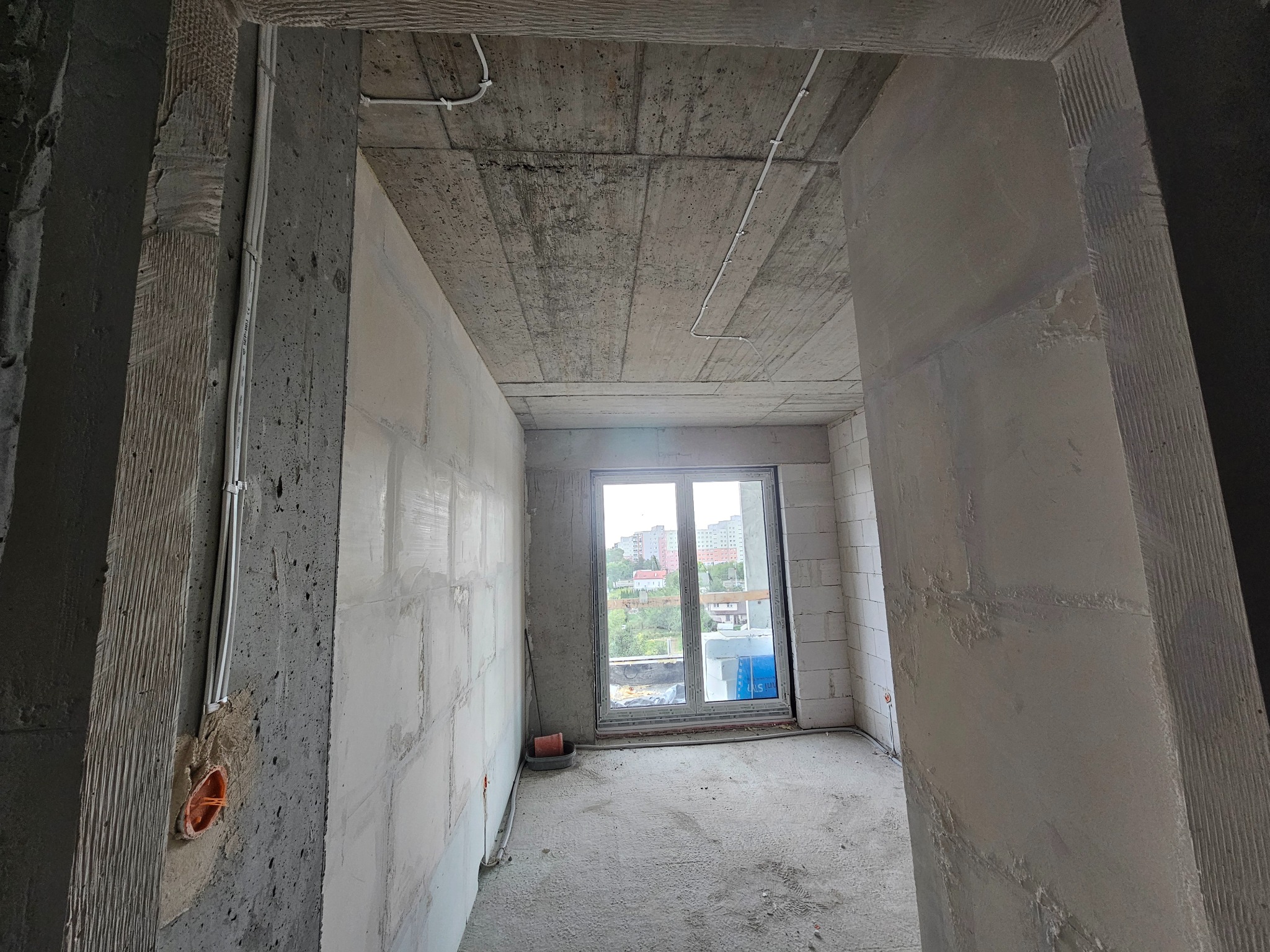 Surowy stan mieszkania w Podzamczu: widoczne przewody elektryczne na suficie i ścianach, ściany z betonu komórkowego, okno z widokiem na zieleń. Przygotowanie do prac wykończeniowych.
