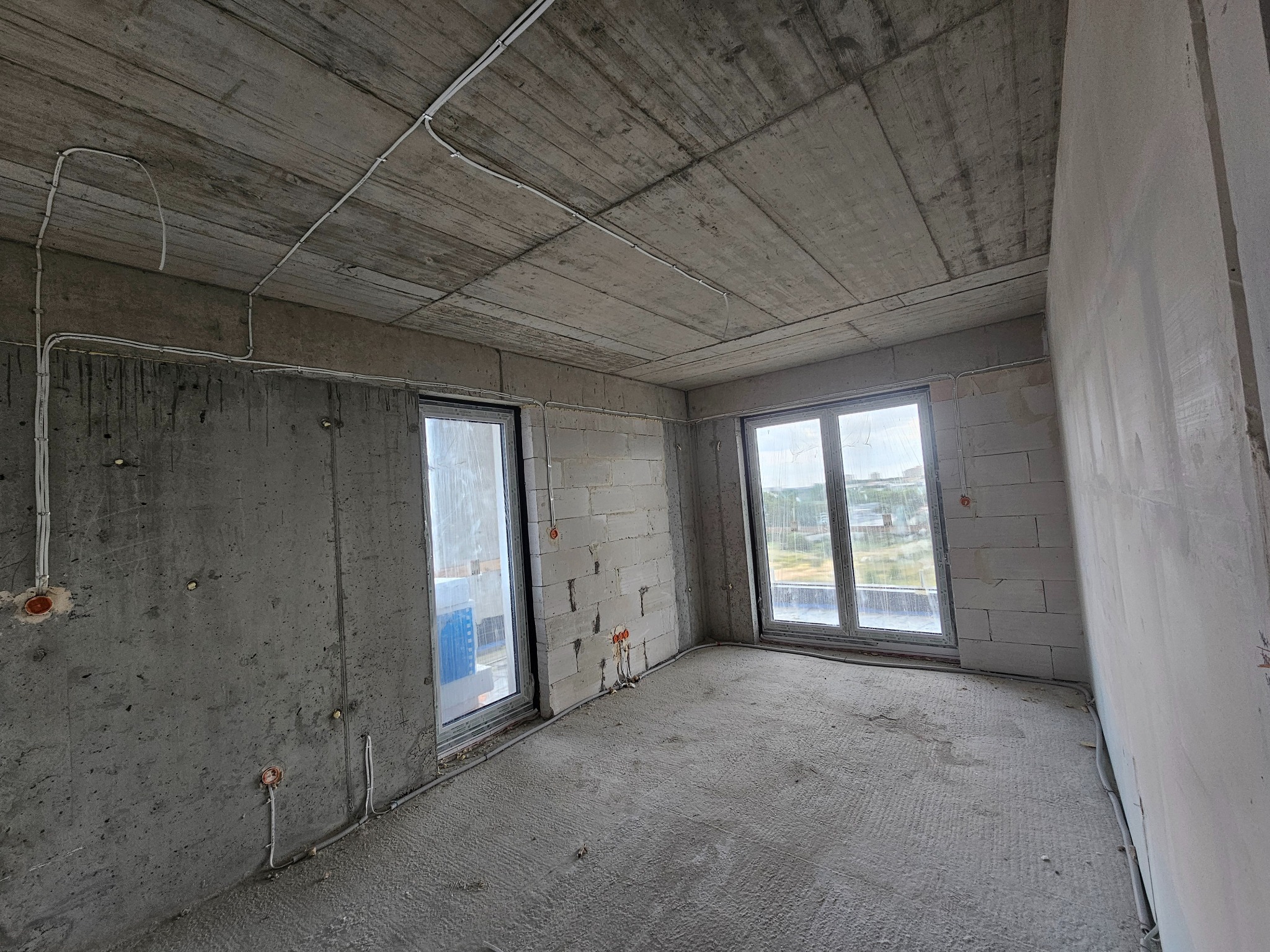 Surowy stan wnętrza: betonowy sufit z instalacją elektryczną, ściany z betonu komórkowego i wylewka. Widoczne okna z widokiem na zewnątrz.