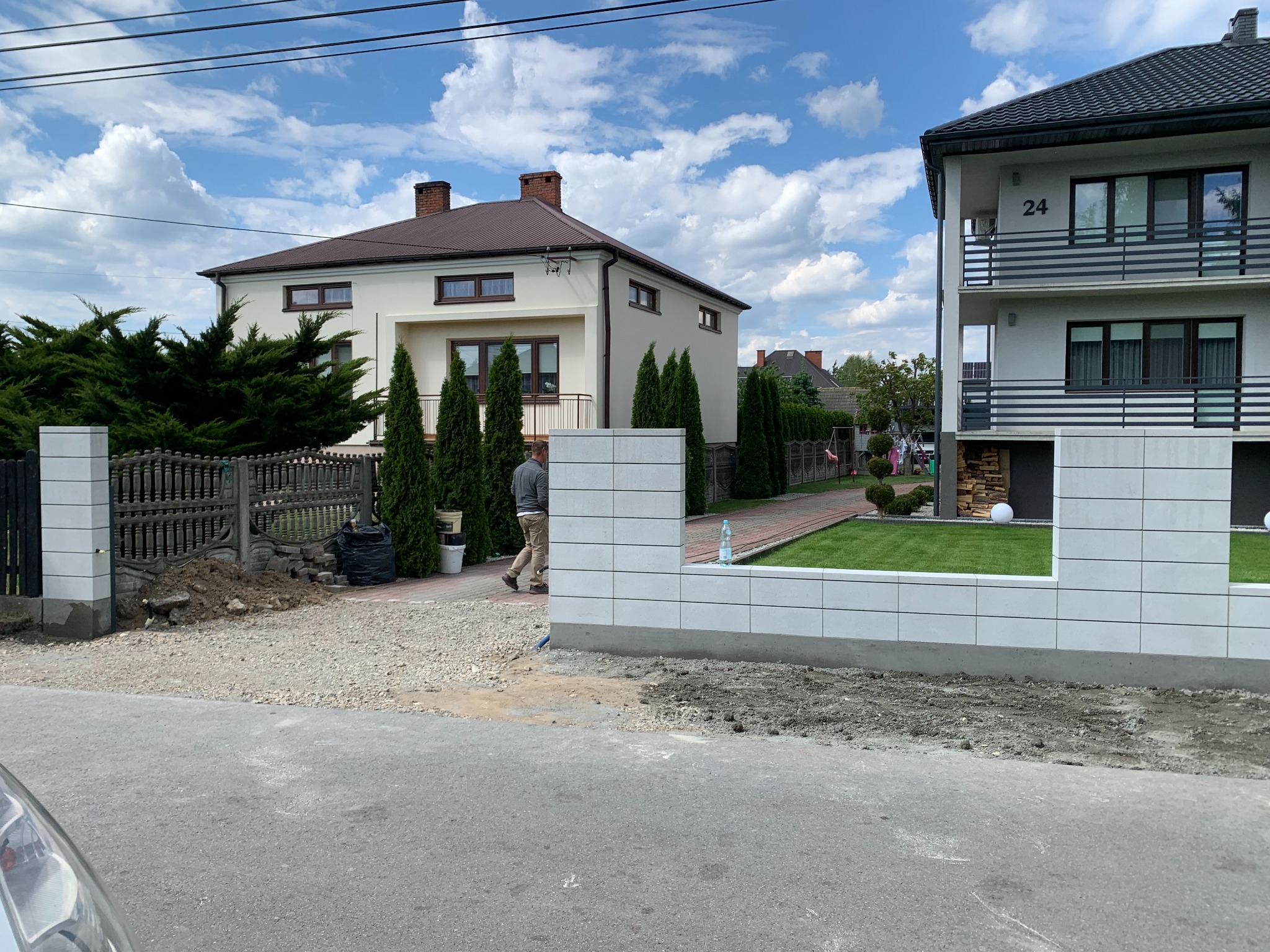 Wznoszenie ogrodzenia z białych bloczków na posesji z dwoma domami. Widoczne elementy starego ogrodzenia i przygotowany teren pod nową konstrukcję. Prace w toku.