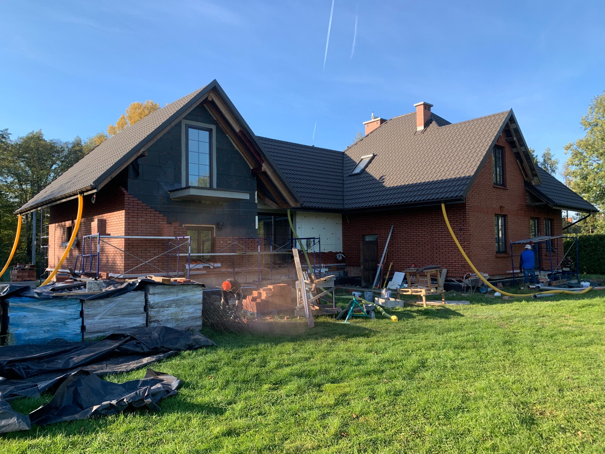 Ceglany dom w trakcie budowy: widoczne rusztowania, materiały budowlane i częściowo ocieplona elewacja. Prace murarskie w toku, z żółtymi wężami wentylacyjnymi.