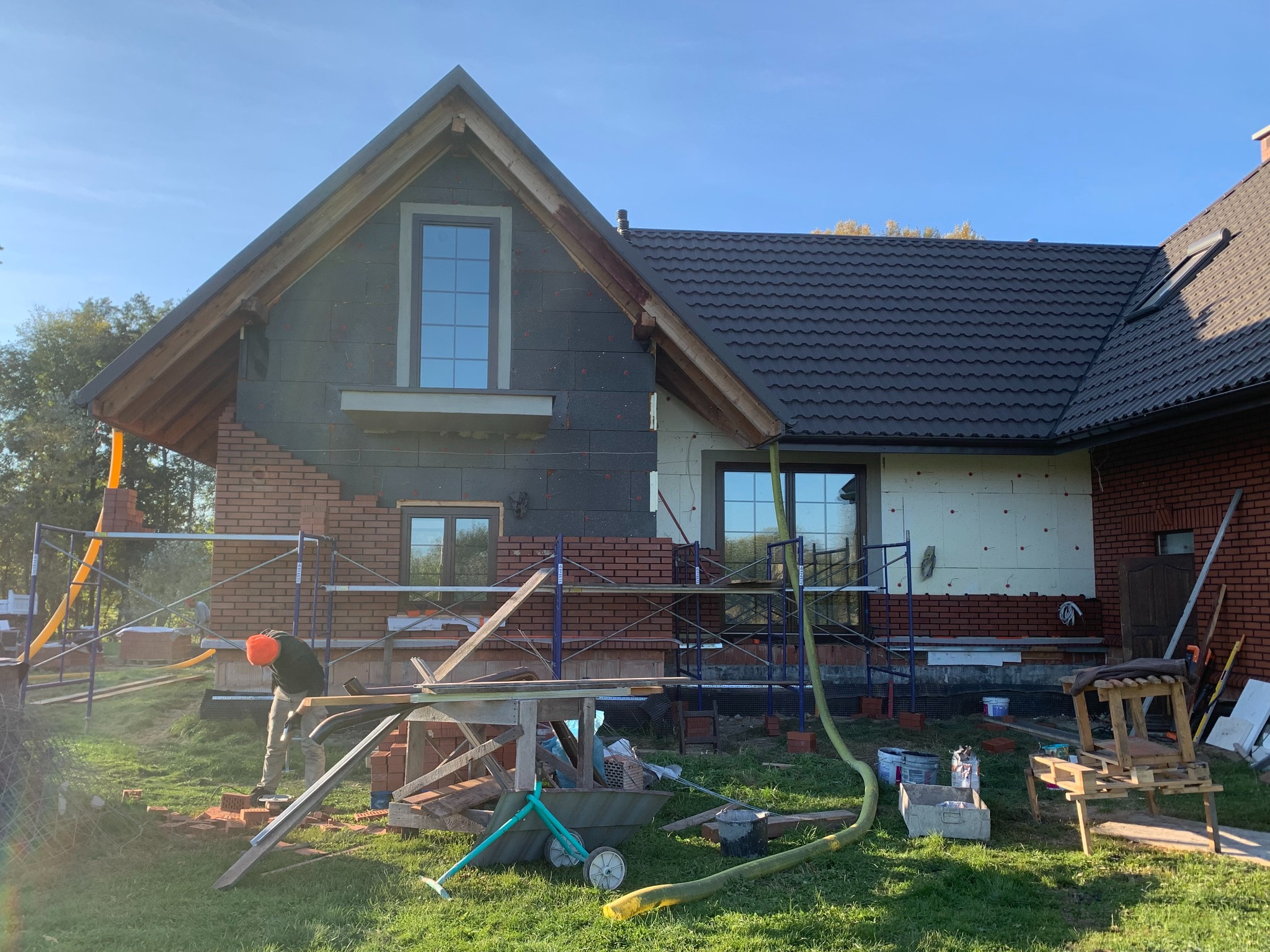 Budowa domu jednorodzinnego: murarz w trakcie kładzenia cegieł na elewacji, widoczne rusztowania, ocieplenie budynku i narzędzia pracy na trawniku.