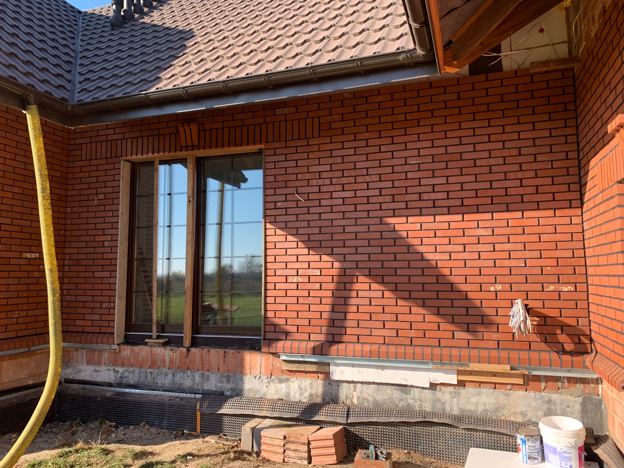 Ceglana ściana domu w trakcie budowy, widoczne okno z drewnianą ramą, żółty wąż i materiały budowlane. Ujęcie z zewnątrz, w słoneczny dzień.