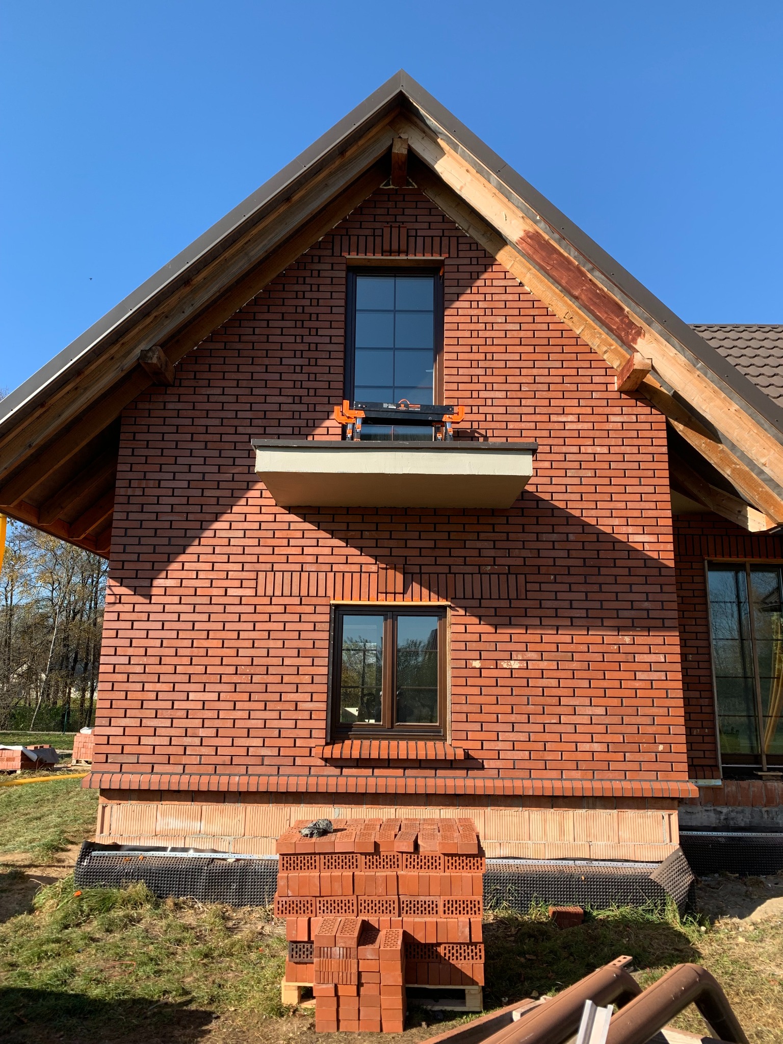 Ceglana ściana domu w trakcie budowy, widoczne okna, drewniana konstrukcja dachu i palety z cegłami. Ujęcie z dołu, w słoneczny dzień.