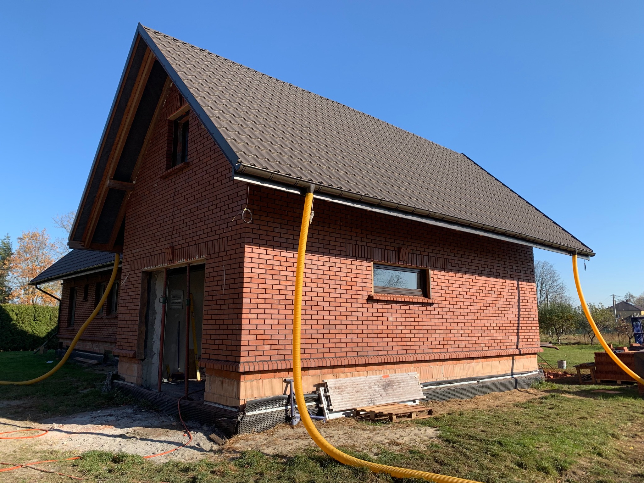 Ceglany dom jednorodzinny z brązowym dachem w trakcie budowy, widoczne żółte rury wentylacyjne, okna i drzwi bez wykończenia, błękitne niebo w tle.