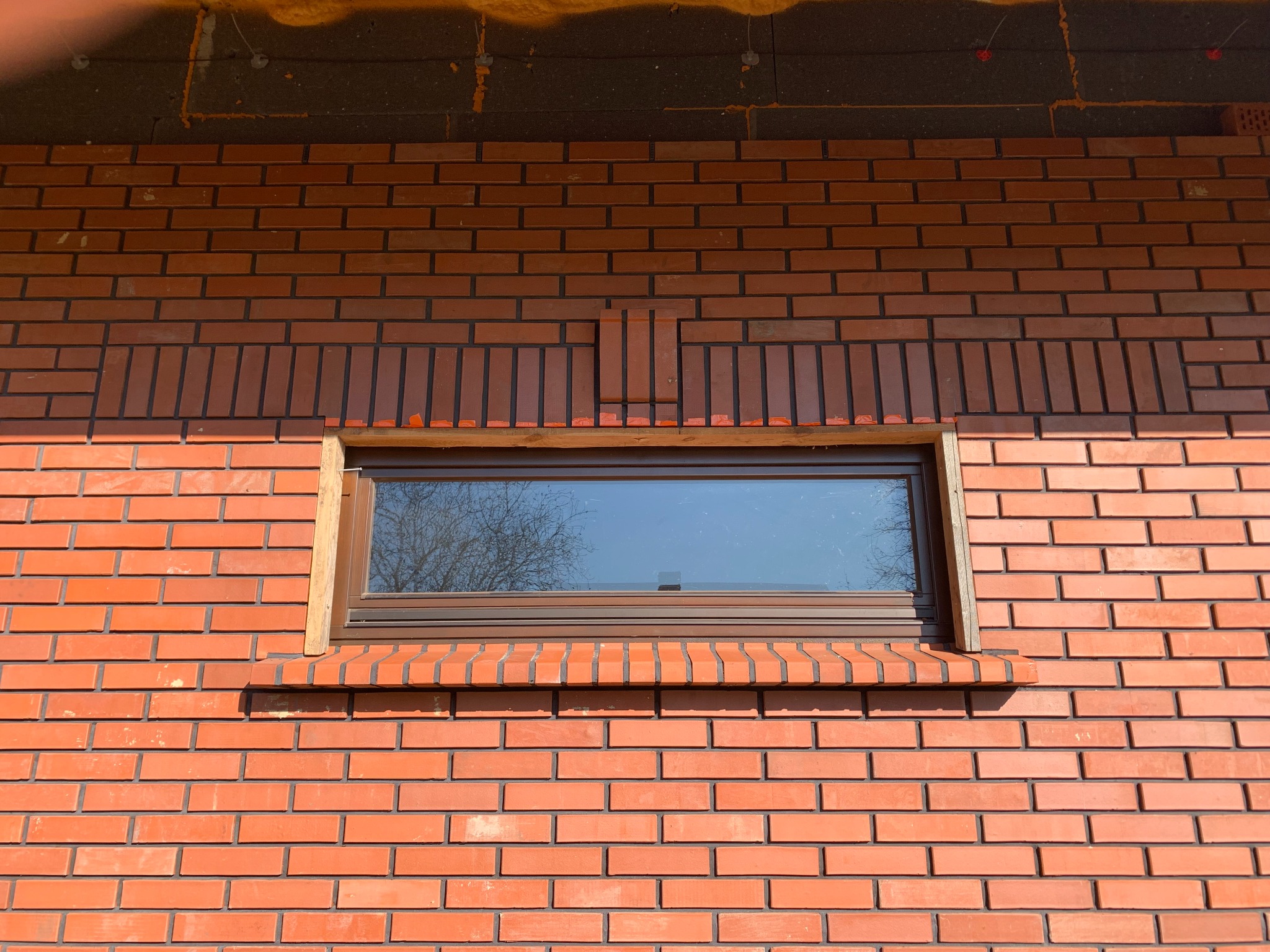 Ceglana ściana domu z oknem, widoczne detale wykończenia wokół okna, brązowe ramy okienne, odbicie drzewa w szybie, fragment ocieplenia na górze.