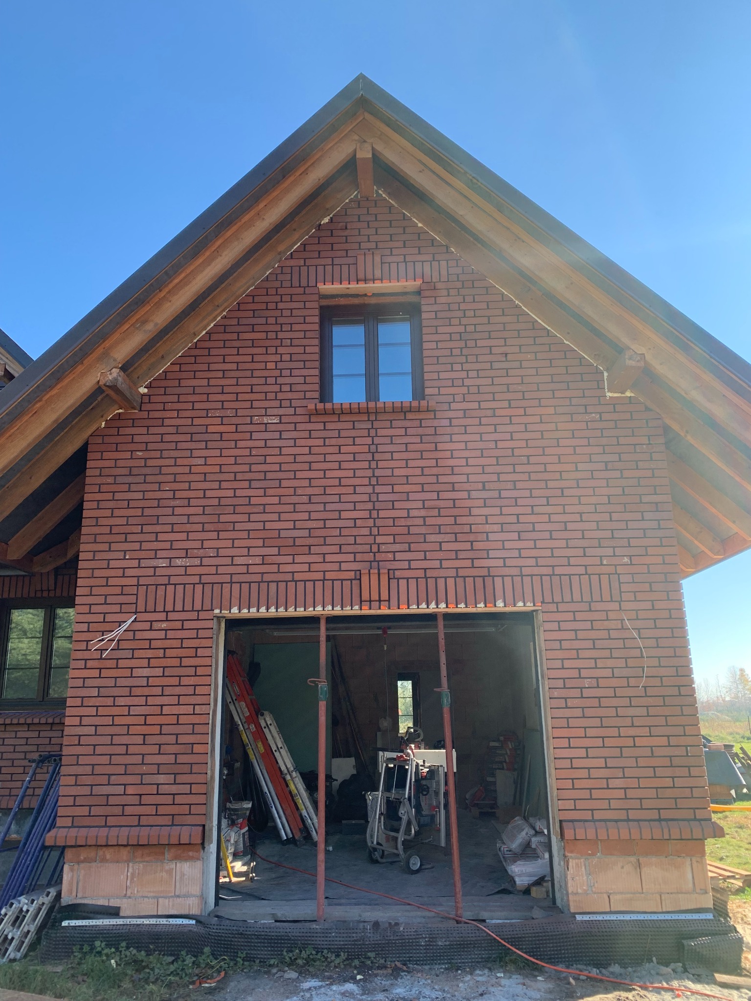 Surowy stan domu z czerwonej cegły z widocznym oknem i otwartym garażem, gdzie widać narzędzia i materiały budowlane. Niebo błękitne w tle.