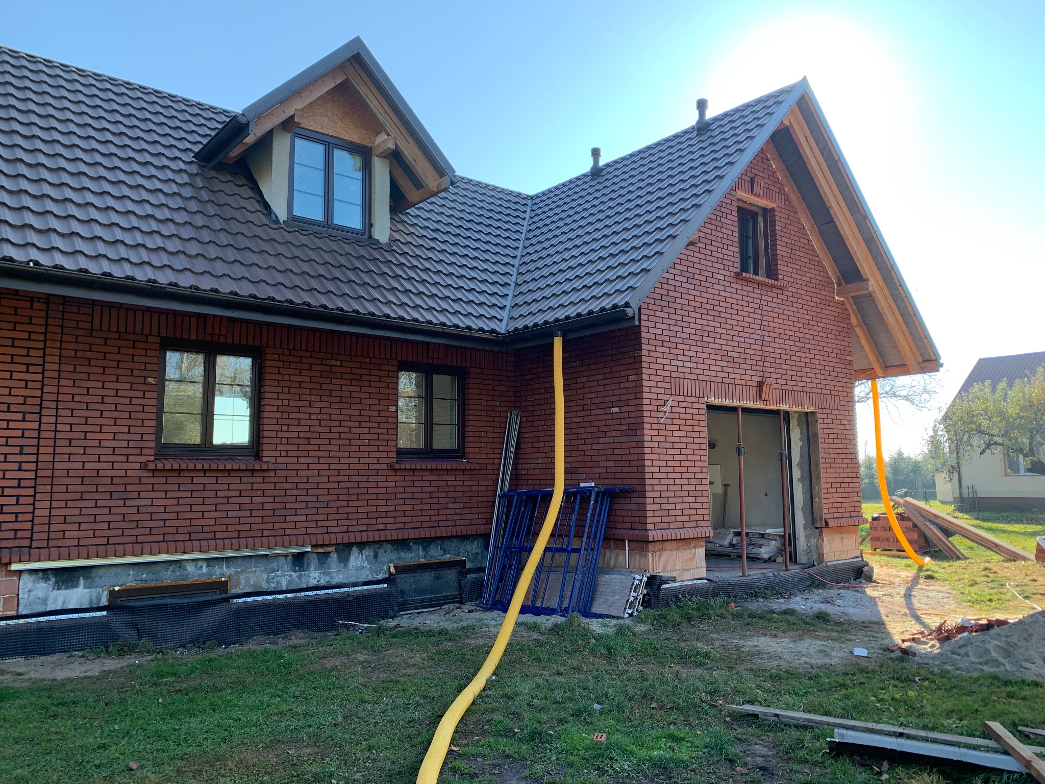Ceglany dom w trakcie budowy, widoczne rusztowania, żółty wąż i materiały budowlane. Dach pokryty brązową dachówką. Słoneczny dzień.