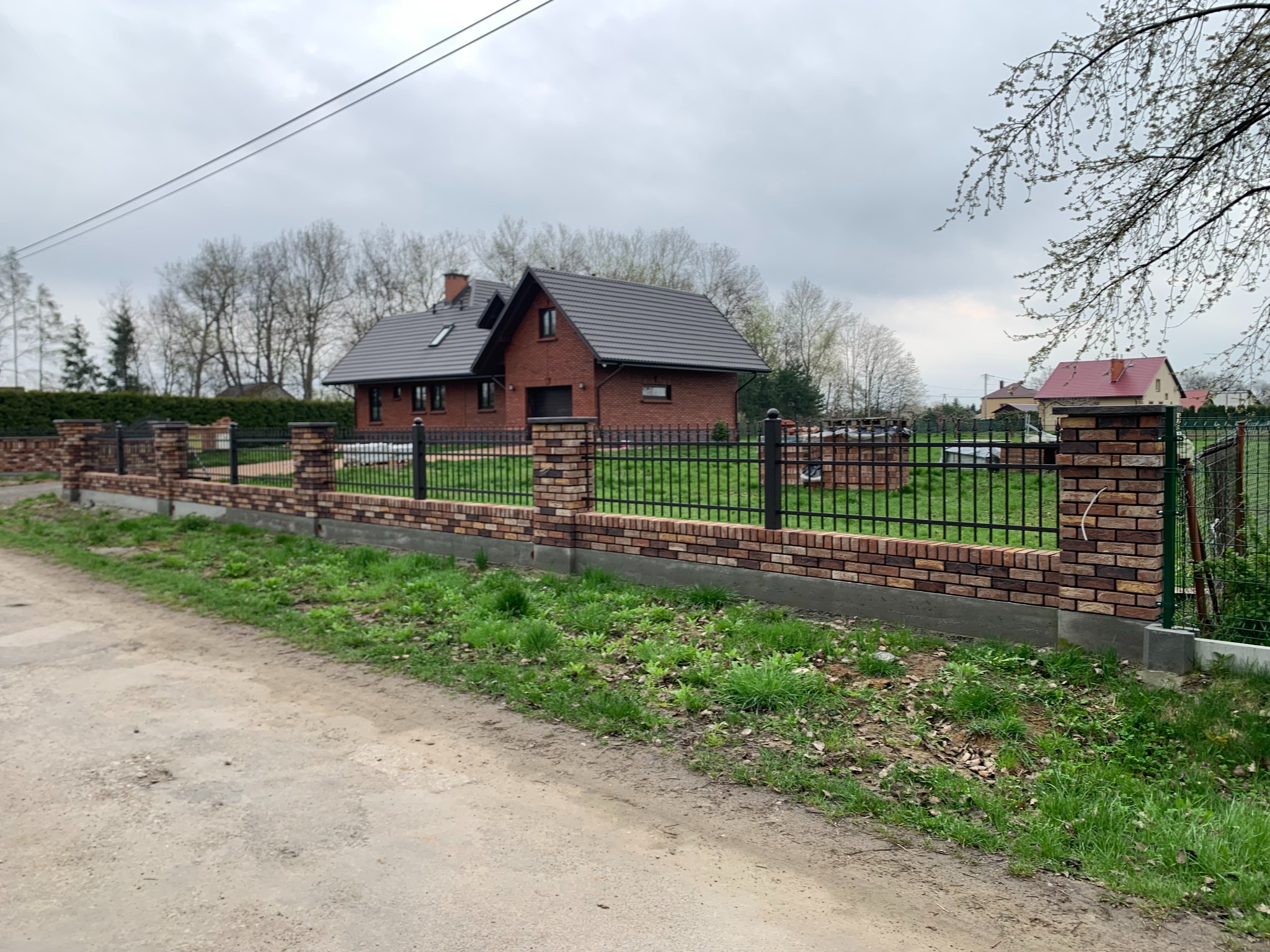 Nowoczesne ogrodzenie z cegły klinkierowej i metalowych przęseł, otaczające posesję z domem w tle. Widok z zewnątrz, w pochmurny dzień.