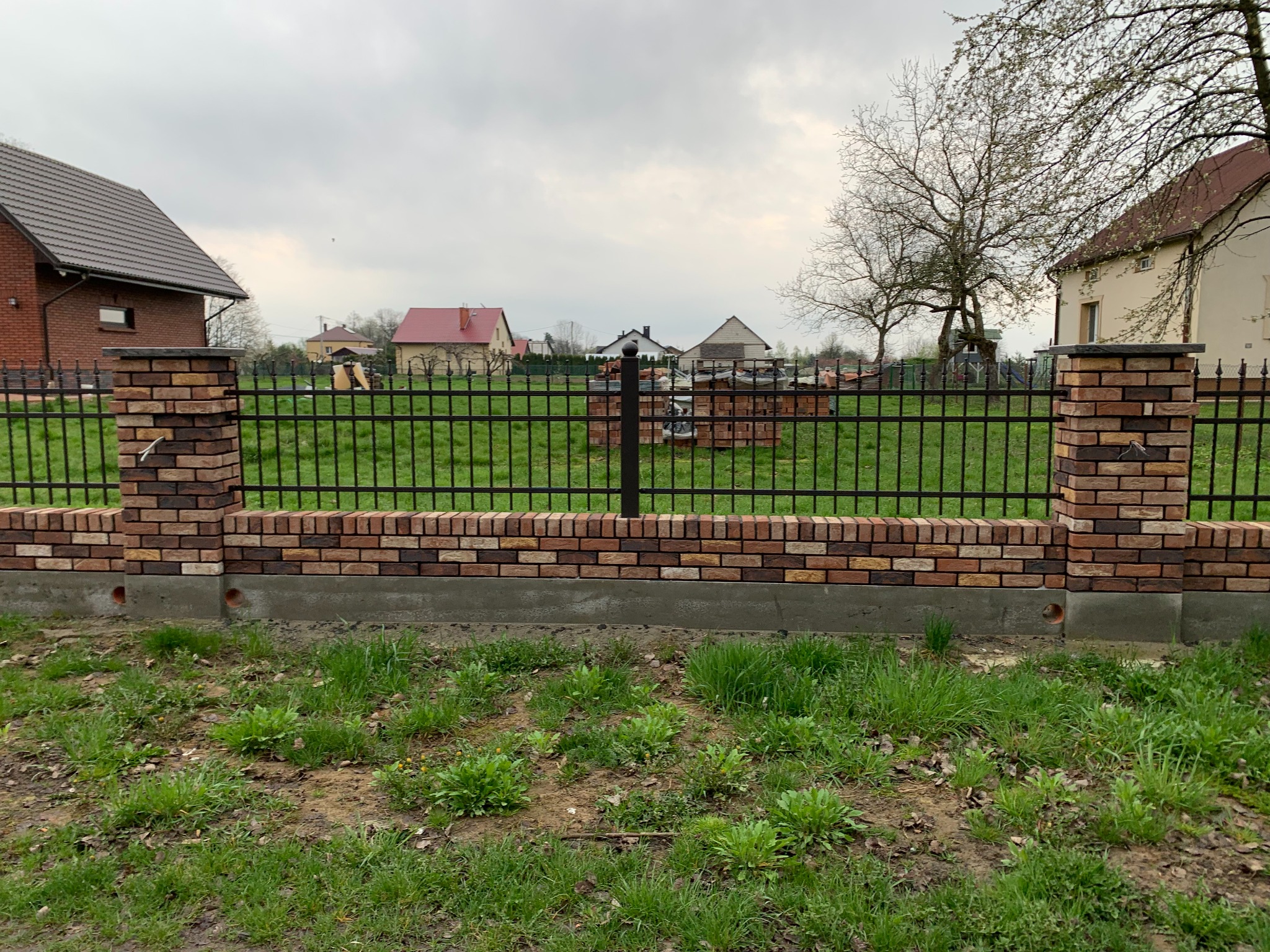 Nowoczesne ogrodzenie z cegły klinkierowej i metalowych przęseł na tle zielonego trawnika i zabudowy jednorodzinnej w pochmurny dzień. Widoczne detale wykończenia.