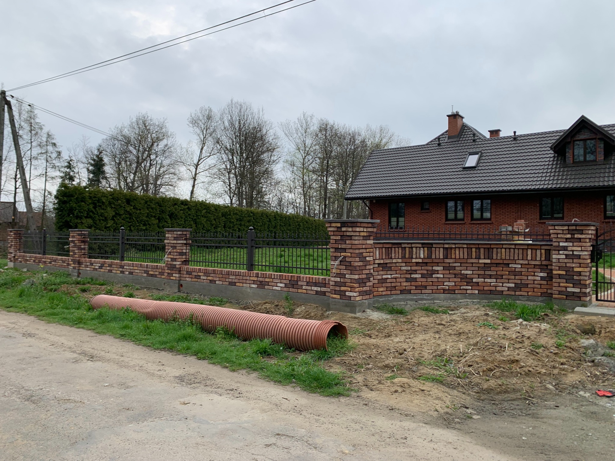 Nowoczesne ogrodzenie z cegły klinkierowej i metalowych paneli na tle domu. Rura kanalizacyjna leży obok, sugerując prace ziemne.