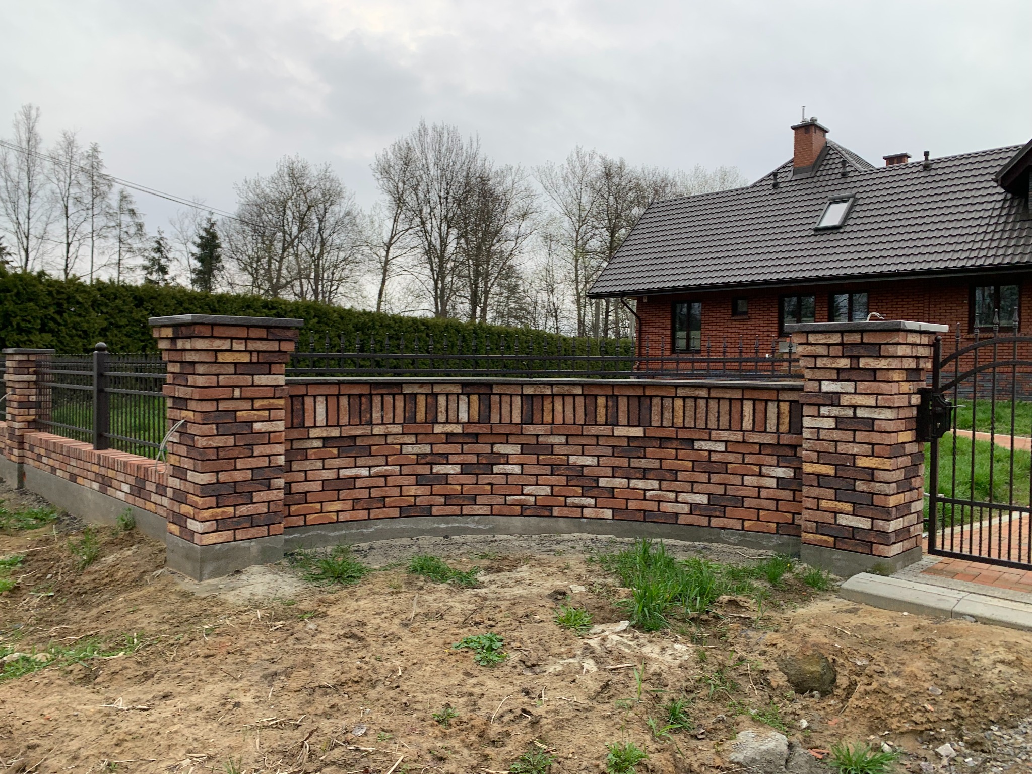 Mur z cegły klinkierowej z metalowym ogrodzeniem na tle domu jednorodzinnego. Nietypowy kształt muru, świeżo posadzona trawa. Elementy wykończeniowe.