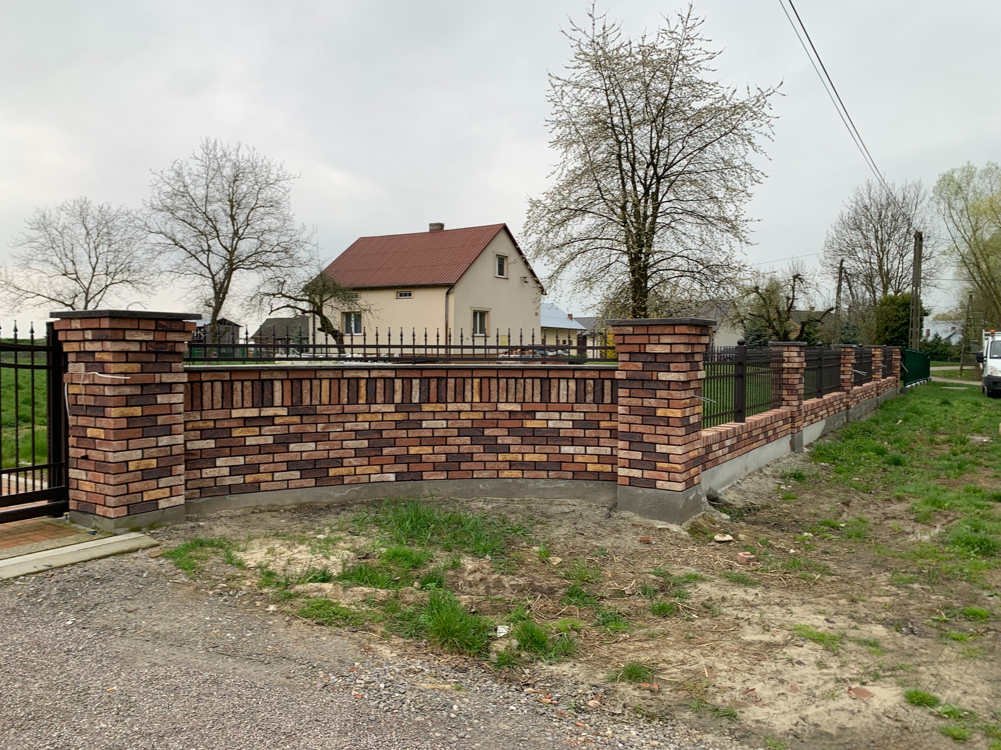 Nowoczesne ogrodzenie z cegły klinkierowej i metalowych paneli w Sokołowie Małopolskim. Solidna konstrukcja, estetyczne wykończenie, widok na dom w tle.
