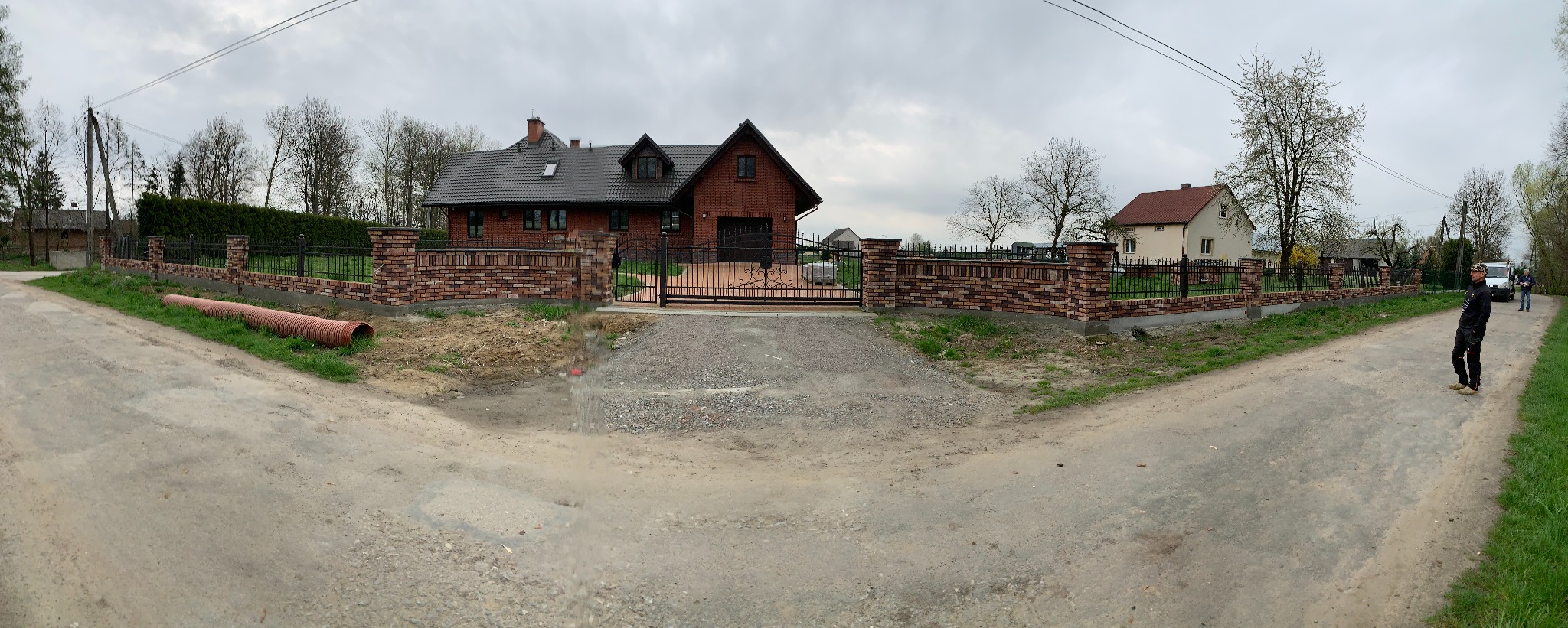 Panorama posesji z ogrodzeniem z cegły klinkierowej i metalowymi przęsłami. Brama wjazdowa z kutego żelaza. W tle dom z czerwonej cegły i szara, pochmurna pogoda.