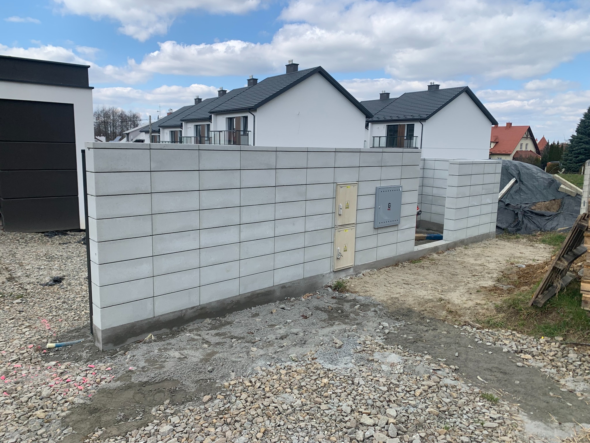 Nowoczesne ogrodzenie z szarej bloczków betonowych, z widocznymi skrzynkami elektrycznymi, na tle szeregu domów z ciemnymi dachami i błękitnym niebem.