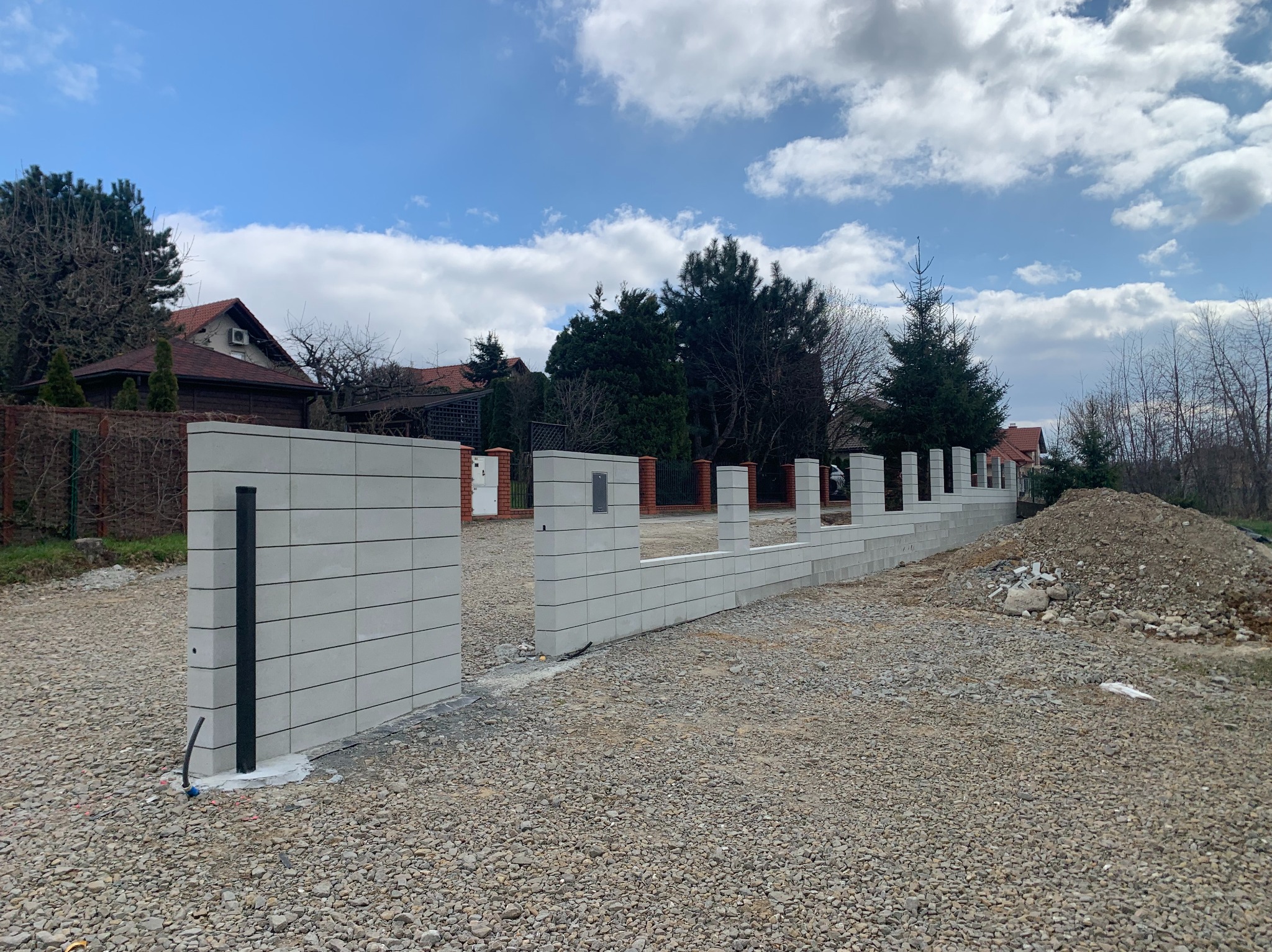 Wznoszony mur z szarej kostki betonowej, tworzący ogrodzenie posesji. Widoczne słupki i fragmenty muru, teren przygotowany pod dalsze prace. W tle domy i drzewa.