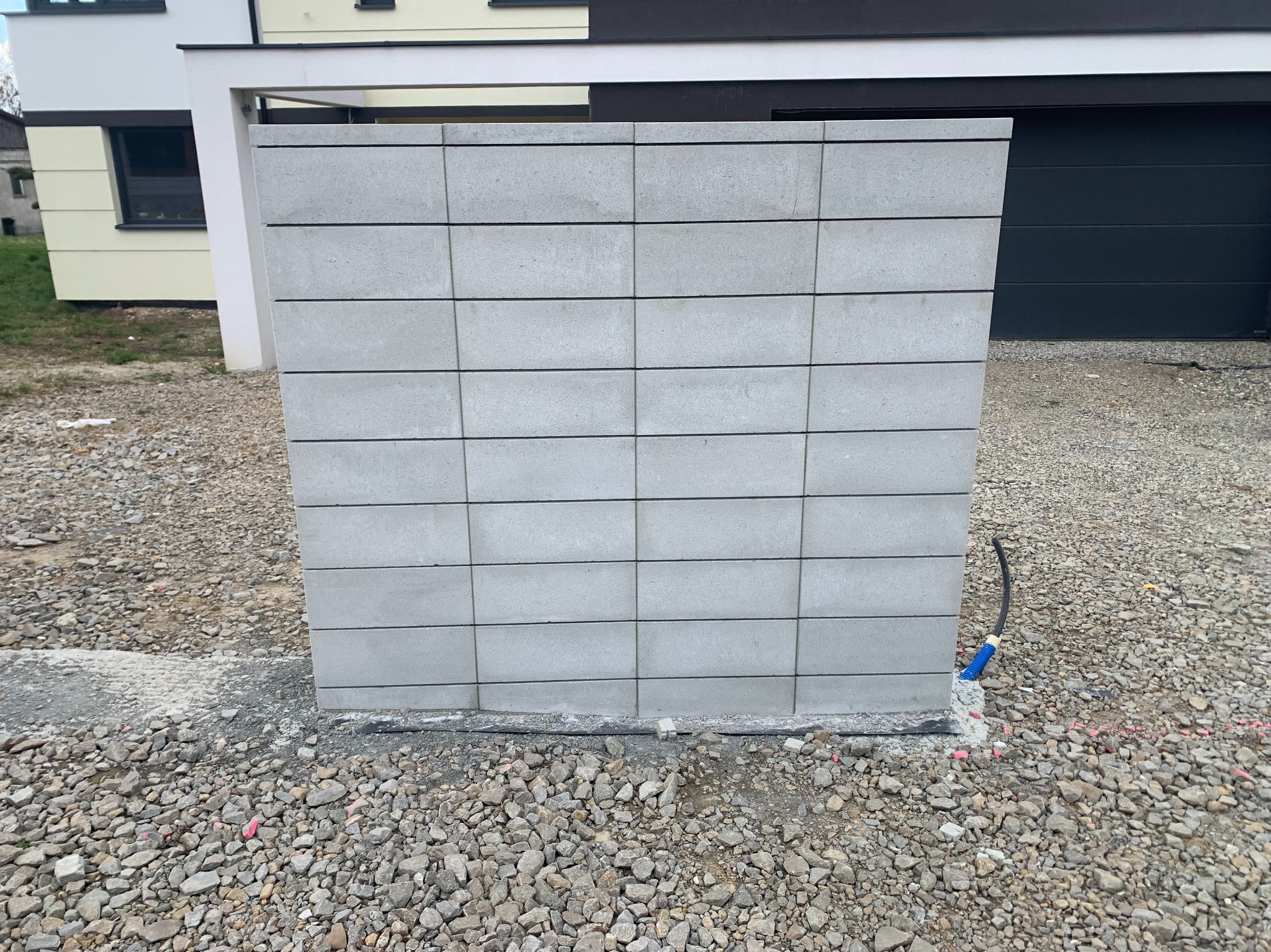 Fragment ogrodzenia z szarej kostki betonowej na żwirowym podłożu, widoczny kabel w peszlu. W tle budynek z garażem.