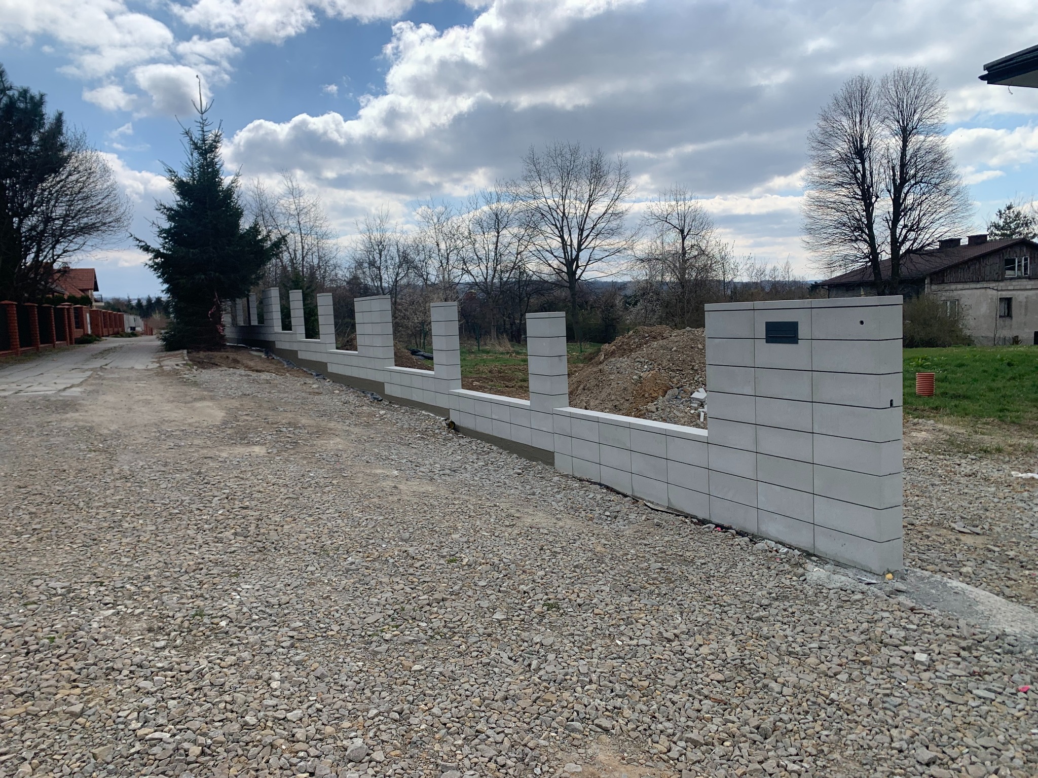 Wznoszenie ogrodzenia z szarej, betonowej kostki modułowej. Widoczne słupki i fragment muru. W tle drzewa i budynki. Grunt utwardzony drobnym kamieniem.