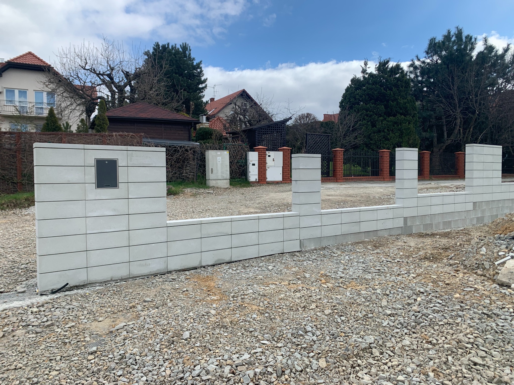 Nowoczesne ogrodzenie modułowe z szarego betonu architektonicznego, z widoczną skrzynką na listy, na tle domów i błękitnego nieba z chmurami.