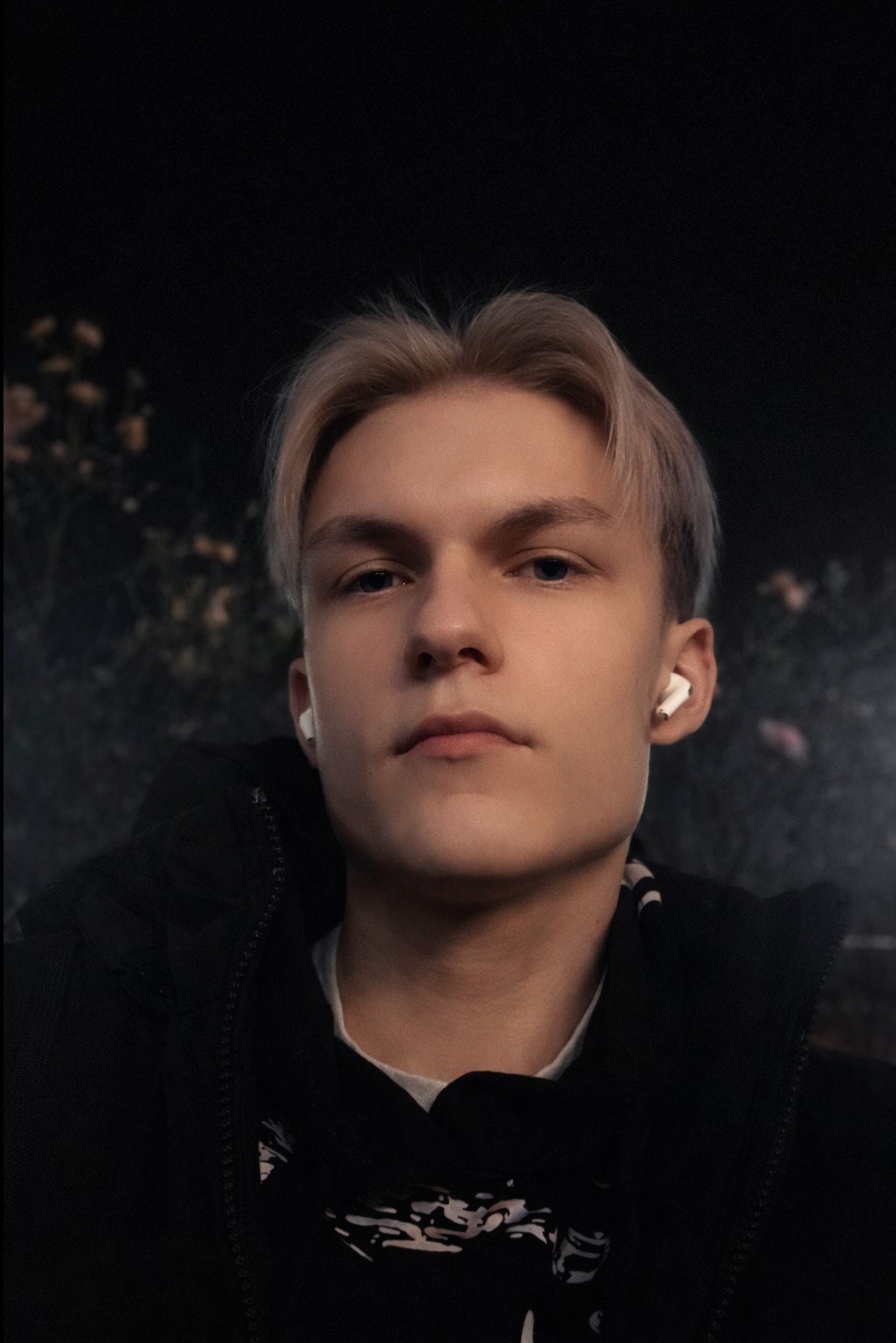 Portret młodego mężczyzny z blond włosami i słuchawkami bezprzewodowymi na tle ciemnego otoczenia z delikatnym oświetleniem.