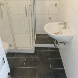 AJS Usługi Ogólnobudowlane Jacek Syktus - Nowoczesna łazienka z kabiną prysznicową, umywalką i ciemną podłogą z płytek. Białe płytki na ścianach. Widoczne elementy instalacji wodnej.