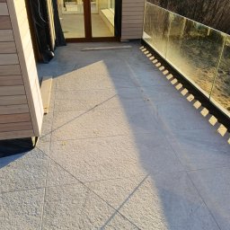 AJS Usługi Ogólnobudowlane Jacek Syktus - Wykończony taras z szarymi płytkami, szklaną balustradą i drewnianą elewacją. Widoczne cienie i detale wykończenia. Ujęcie z góry.