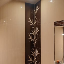 AJS Usługi Ogólnobudowlane Jacek Syktus - Beżowe, błyszczące płytki w łazience z dekoracyjnym, pionowym pasem z motywem bambusa. W tle widoczne lustro z brązową mozaiką.
