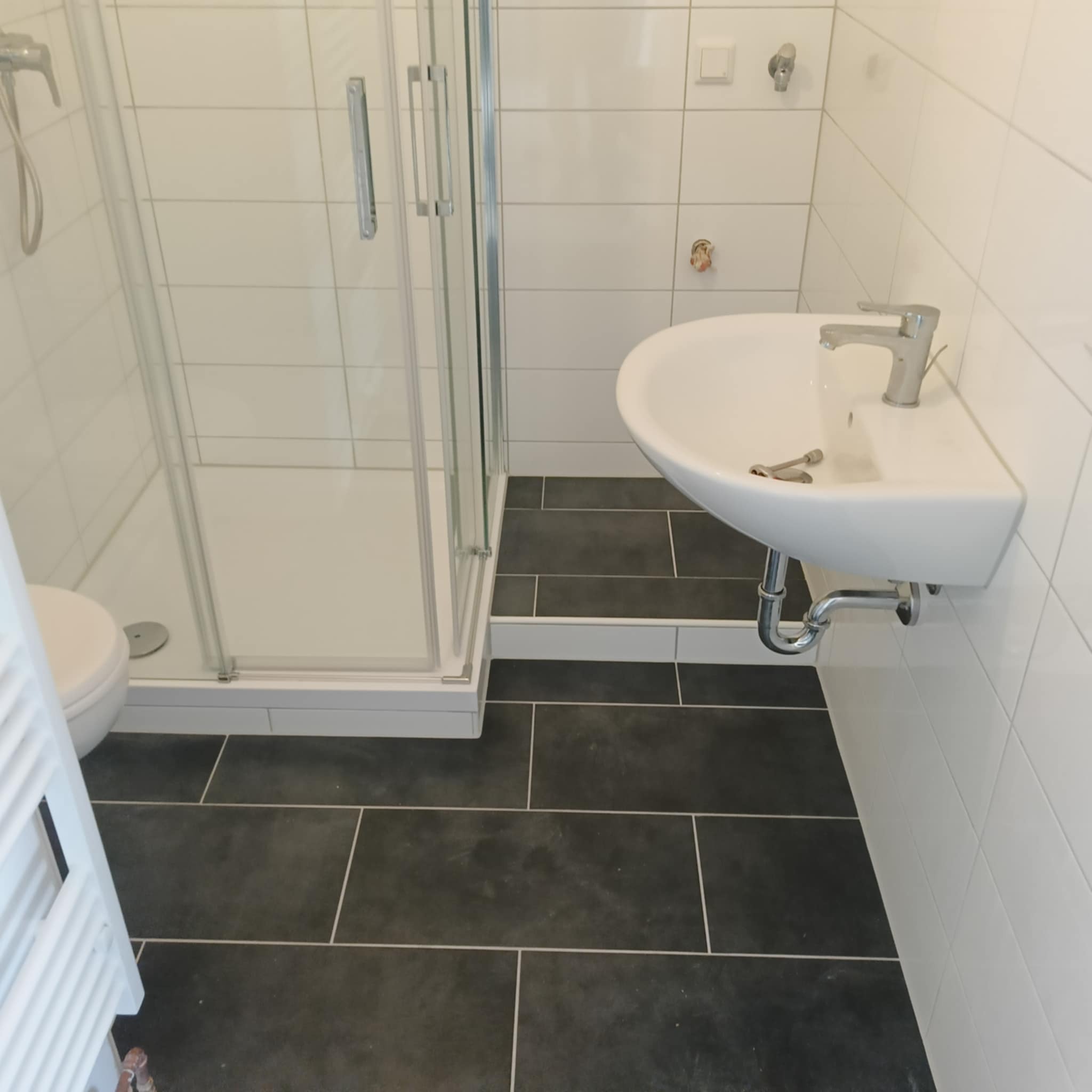 Nowoczesna łazienka z kabiną prysznicową, umywalką i ciemną podłogą z płytek. Białe płytki na ścianach. Widoczne elementy instalacji wodnej.