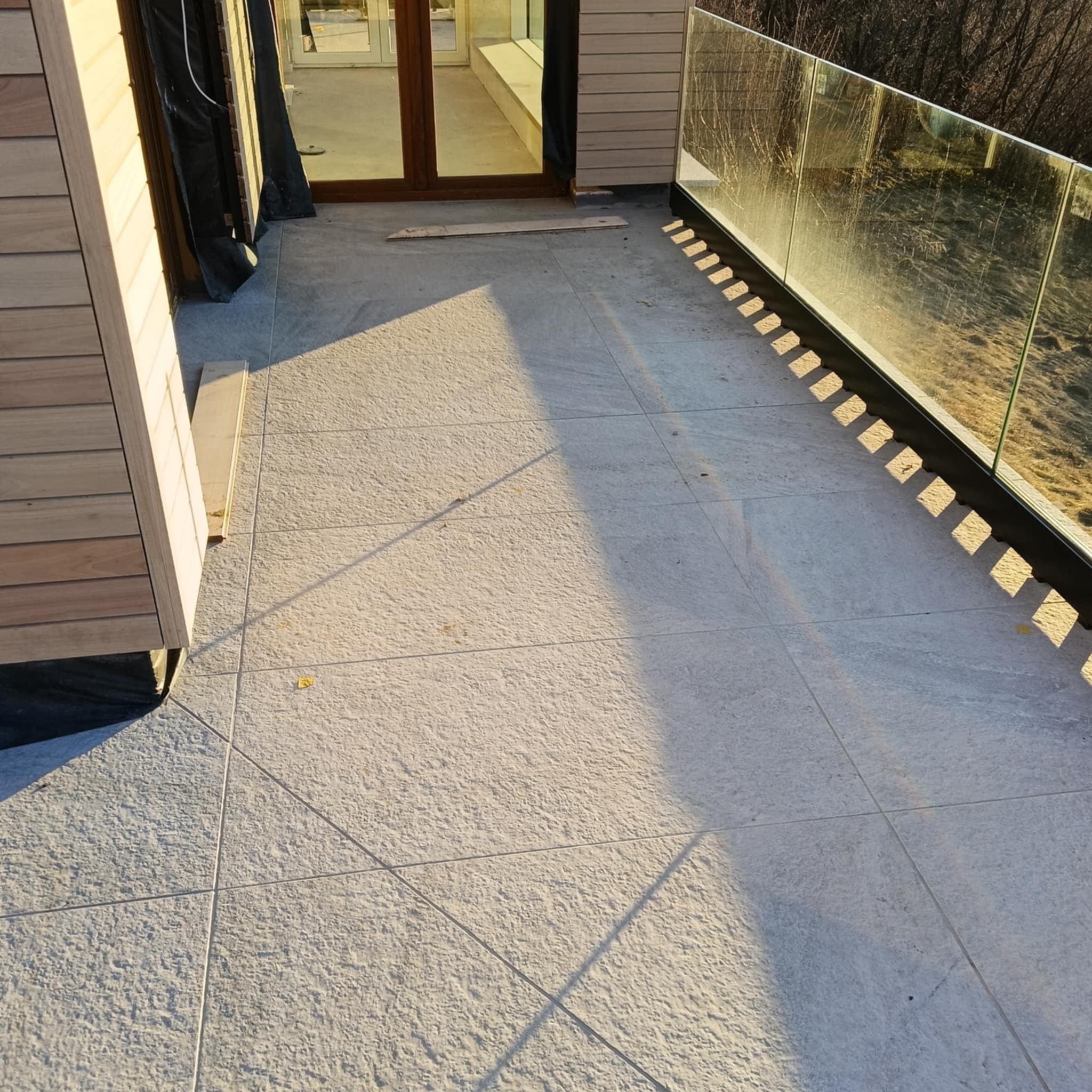 Wykończony taras z szarymi płytkami, szklaną balustradą i drewnianą elewacją. Widoczne cienie i detale wykończenia. Ujęcie z góry.