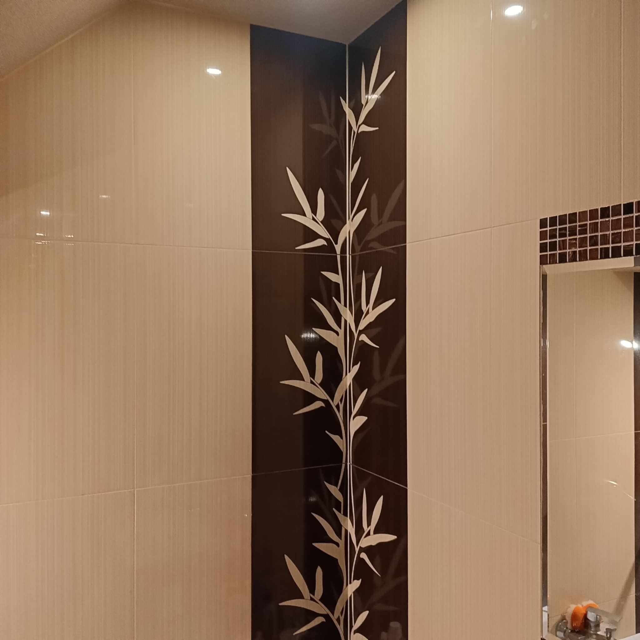 Beżowe, błyszczące płytki w łazience z dekoracyjnym, pionowym pasem z motywem bambusa. W tle widoczne lustro z brązową mozaiką.