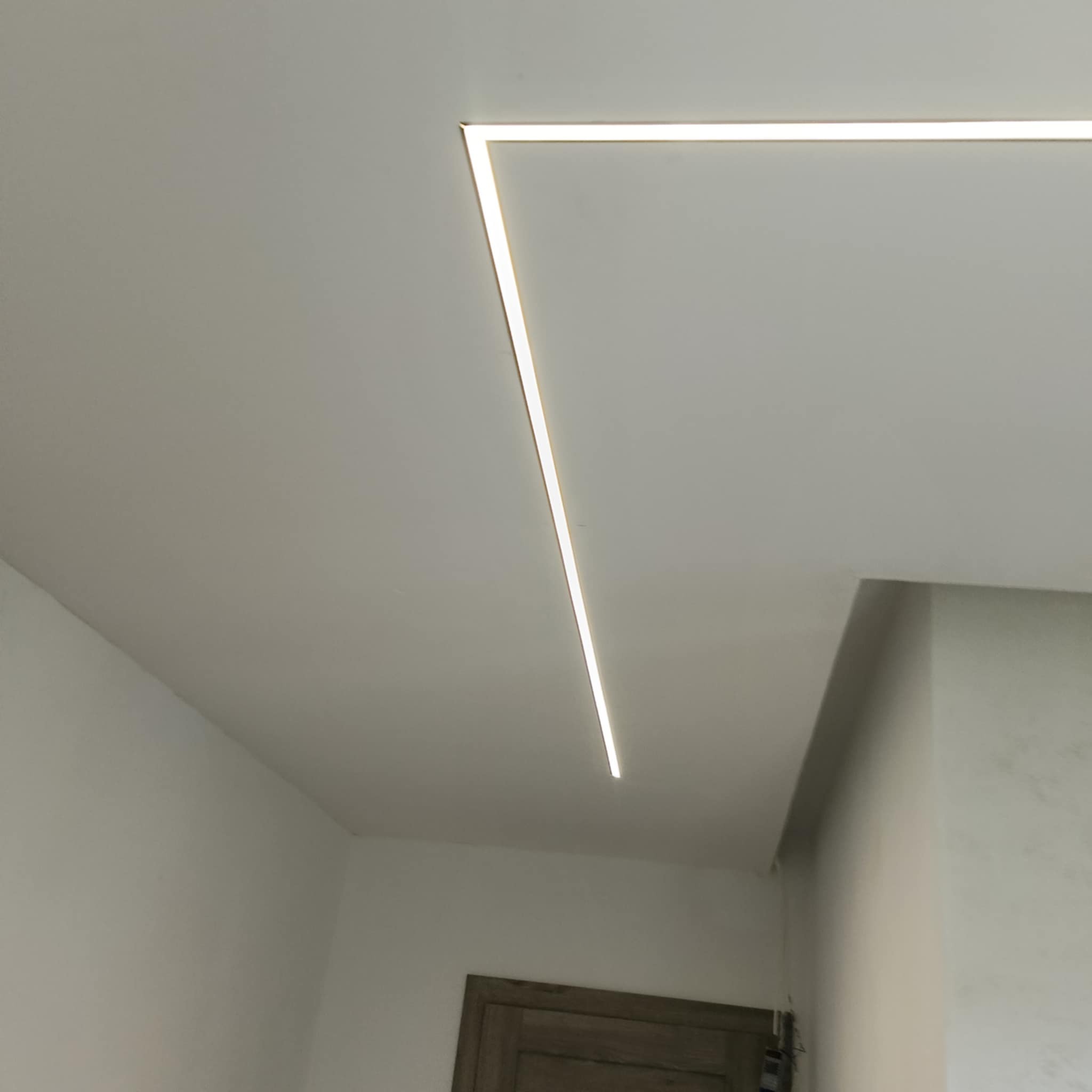 Minimalistyczne oświetlenie LED w kształcie litery L, wkomponowane w sufit podwieszany, tworzące nowoczesny akcent w jasnym wnętrzu.