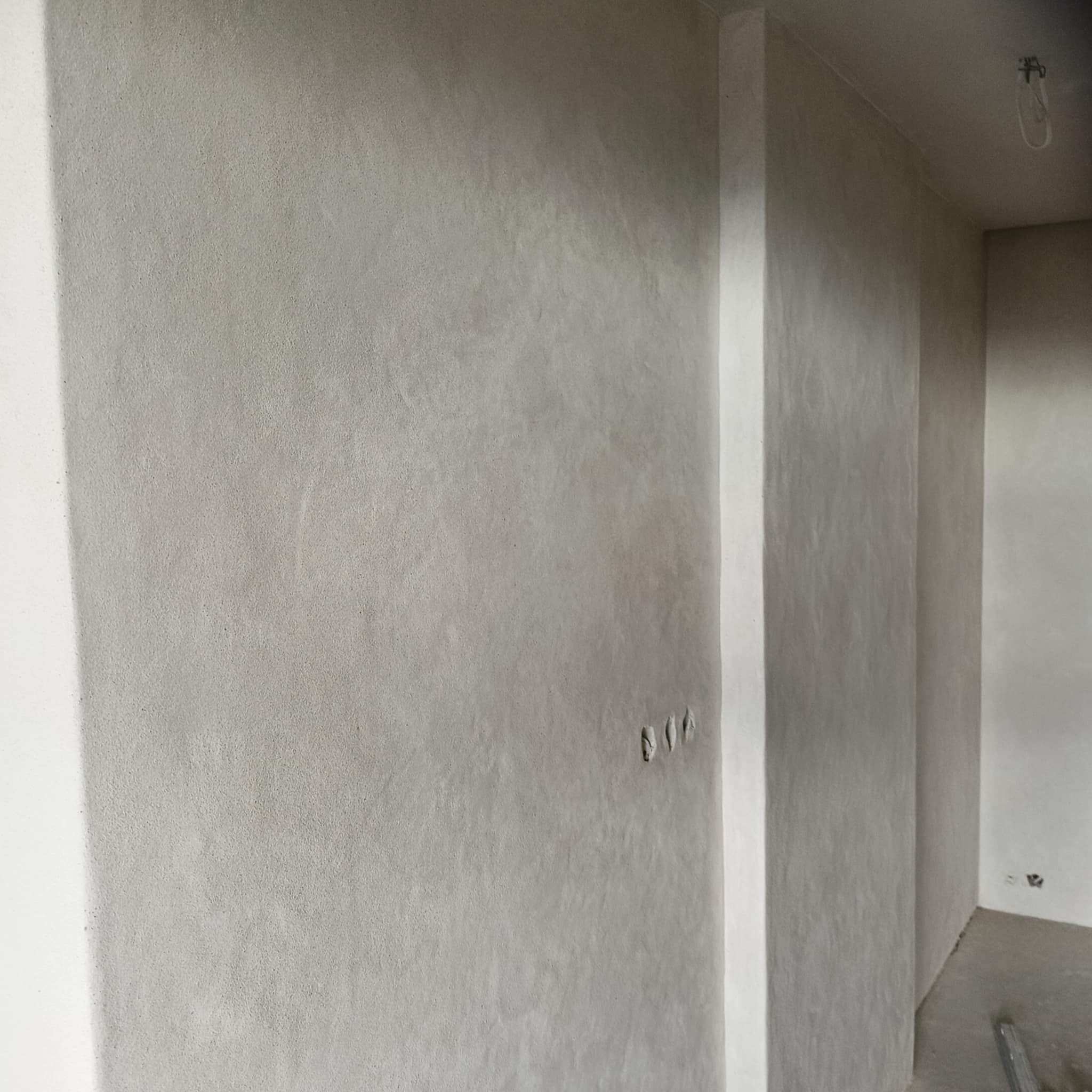 Ściana po szpachlowaniu z widocznymi trzema otworami na gniazdka elektryczne. Szara, gładka powierzchnia w surowym wnętrzu. Widoczny fragment sufitu z żarówką.