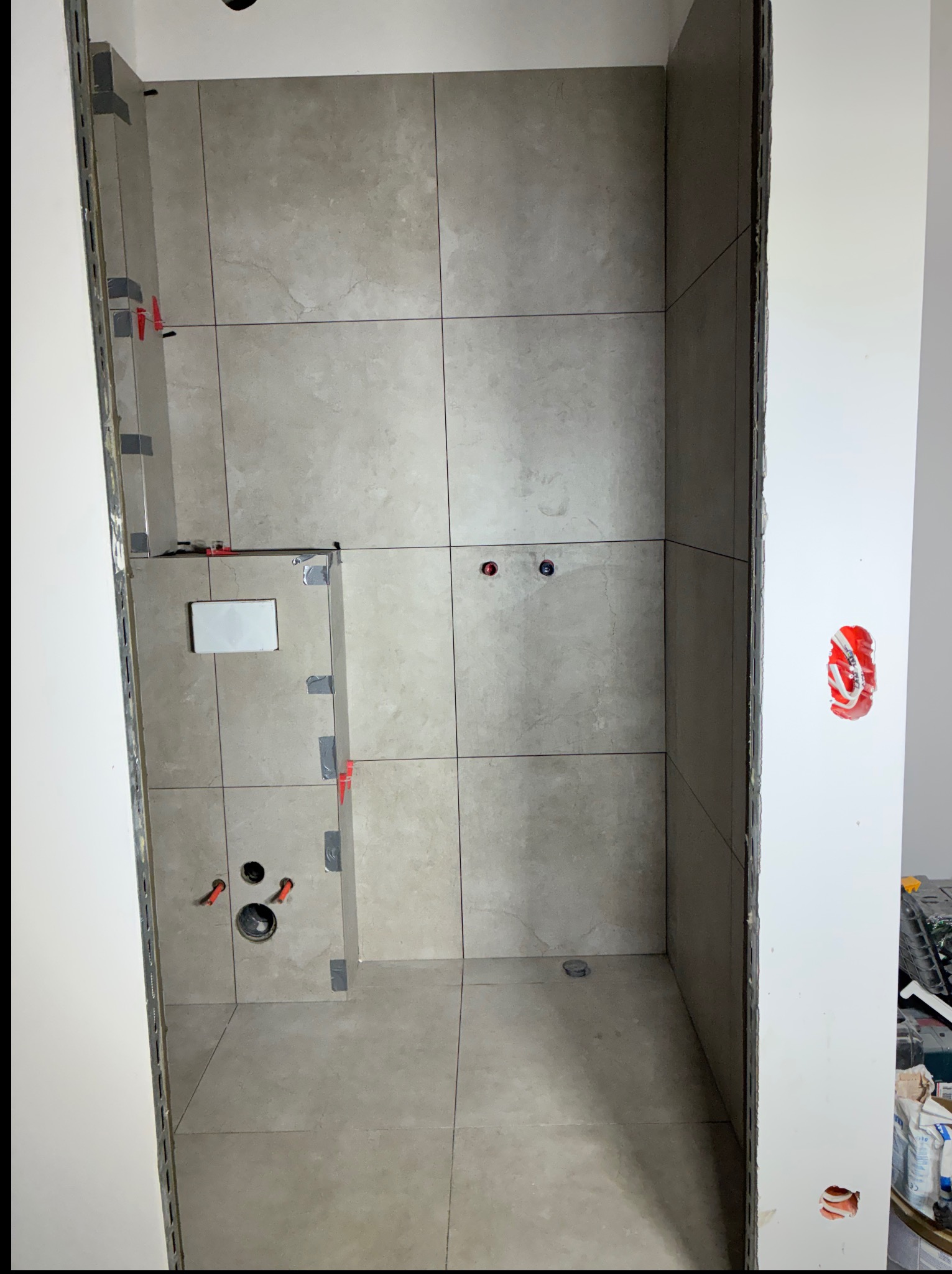 Wnętrze łazienki w trakcie prac wykończeniowych: ułożone płytki w odcieniach szarości, widoczne elementy instalacji i narzędzia. Świeżo położona glazura z krzyżykami dystansowymi.