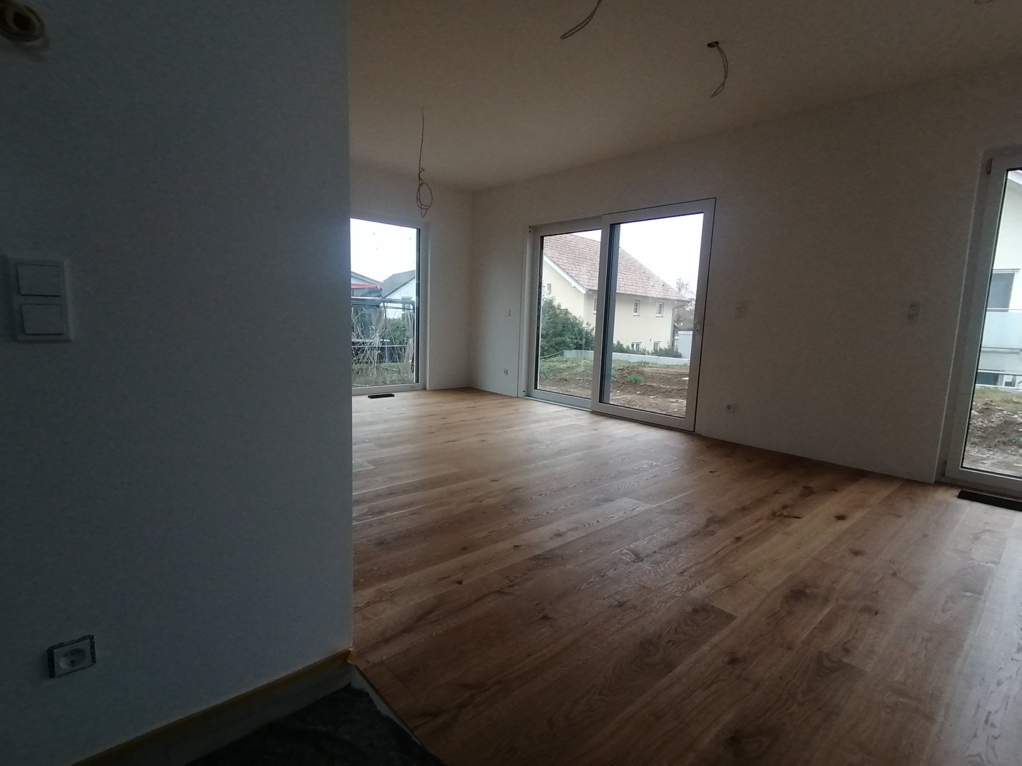 Jasne, przestronne wnętrze z drewnianą podłogą i dużymi oknami balkonowymi. Widoczne gładkie, białe ściany i wystające kable elektryczne. Minimalistyczny styl wykończenia.