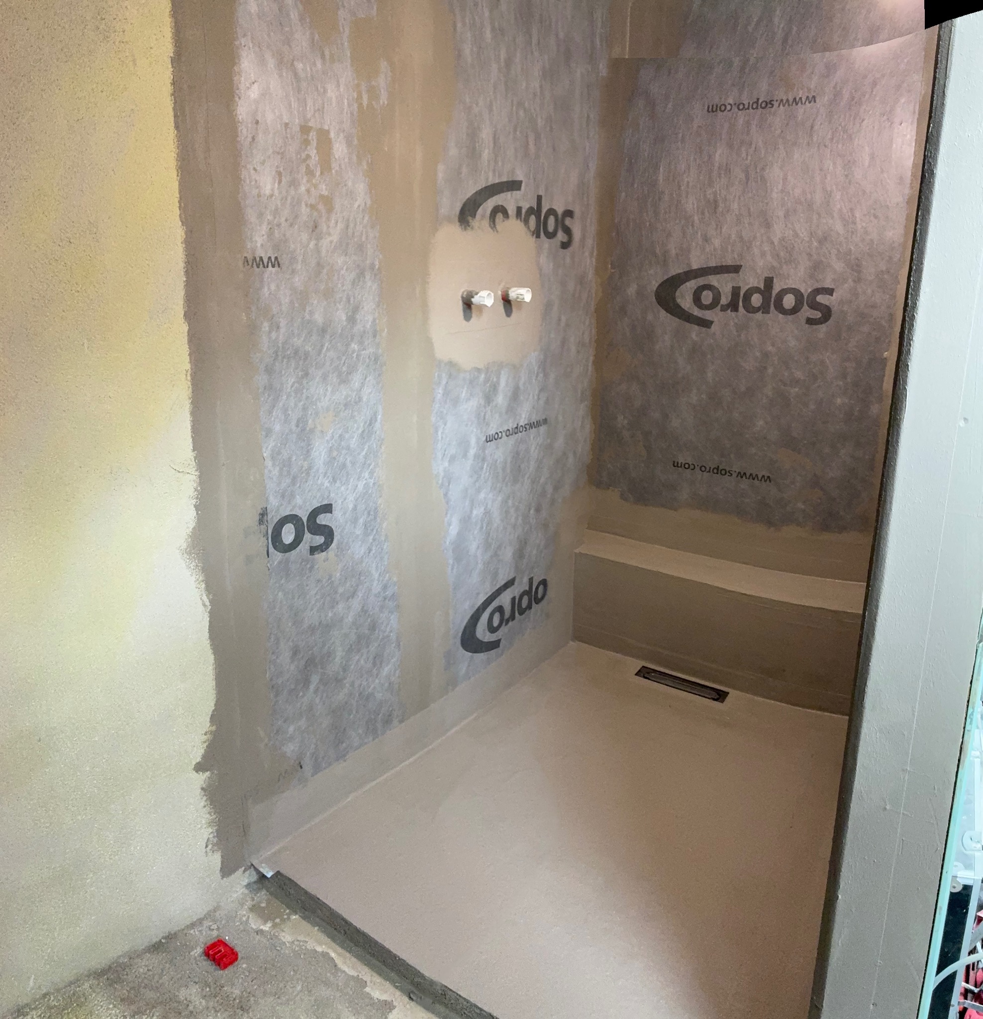 Przygotowanie do hydroizolacji w łazience: ściany zabezpieczone folią w płynie z widocznym logo Cordos, w tle fragment brodzika z odpływem liniowym.