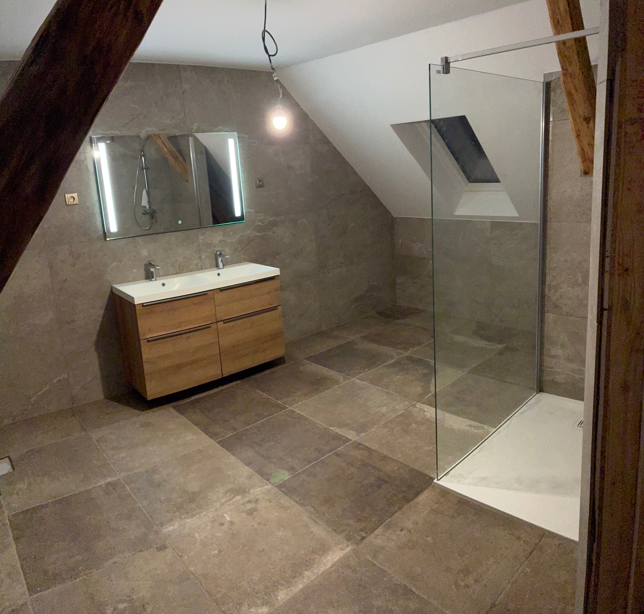 Wykończona łazienka na poddaszu z kabiną prysznicową, umywalką z szafką z jasnego drewna i lustrem z oświetleniem LED. Uwagę przykuwają płytki w odcieniach szarości i drewniane belki.