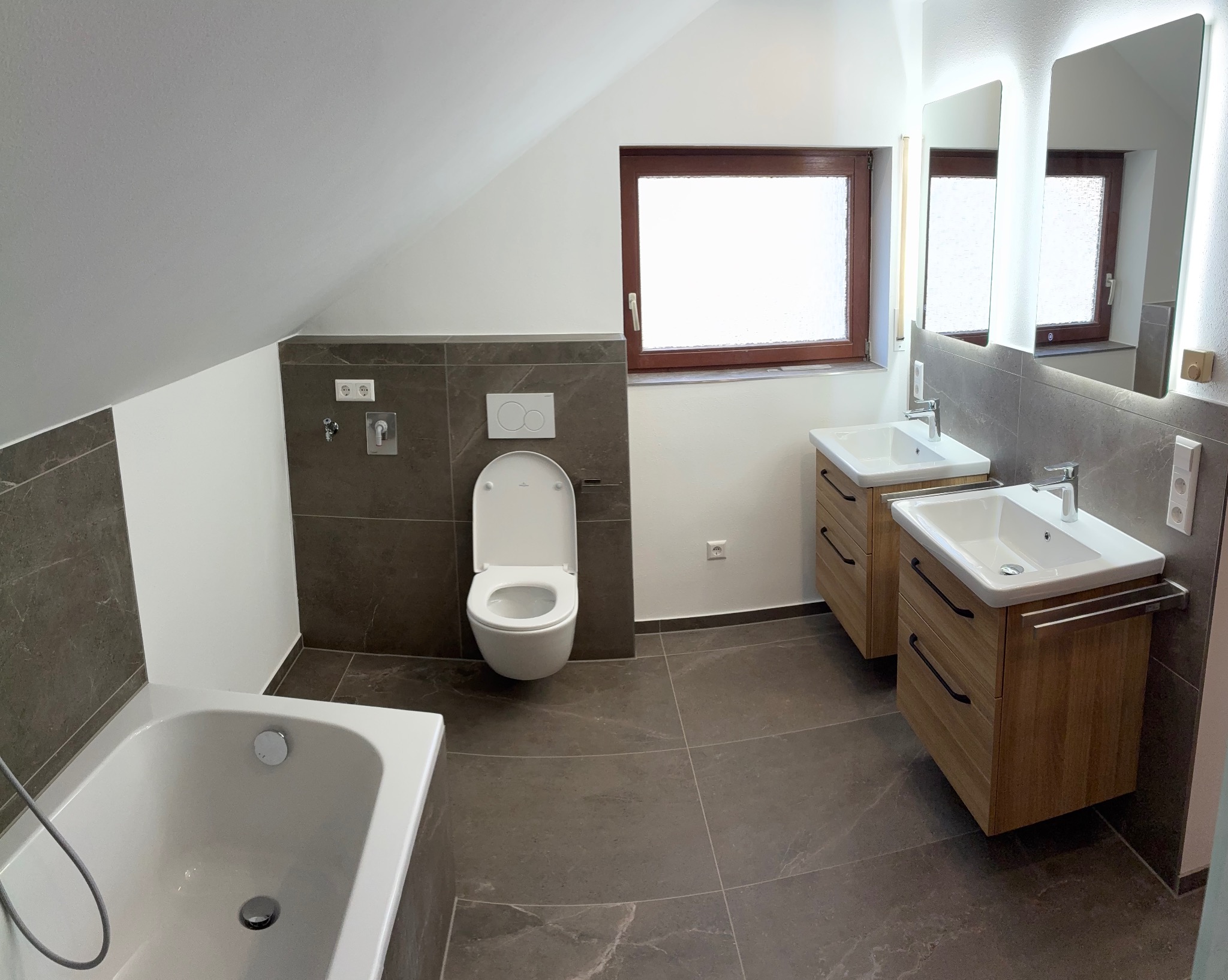 Nowoczesna łazienka na poddaszu z brązowymi płytkami, drewnianymi szafkami pod umywalki, podświetlanymi lustrami i oknem. Uwagę zwraca skośny sufit.