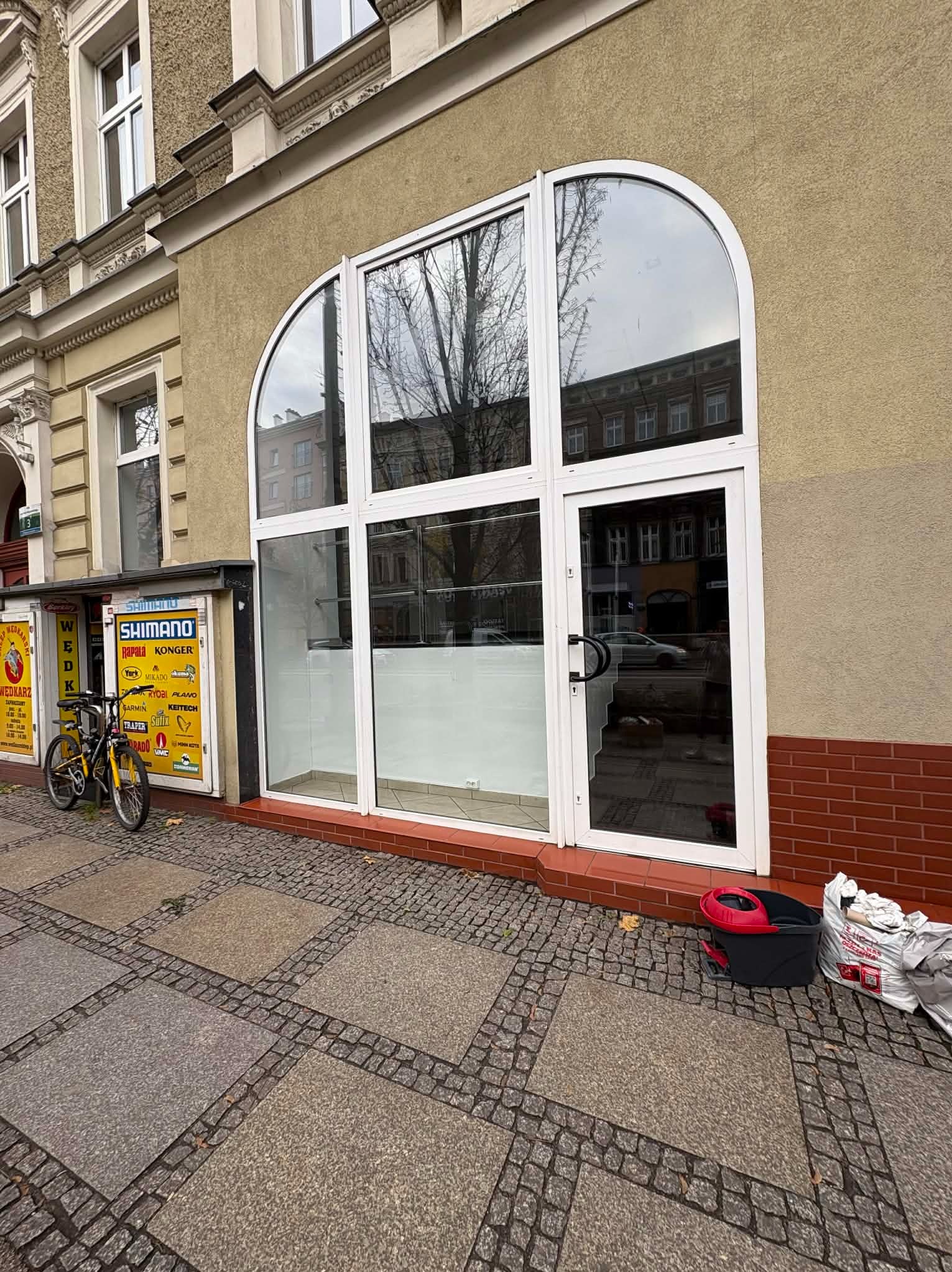 Wnętrze lokalu po sprzątaniu, widoczne przez duże okno z białymi ramami. Na zewnątrz rower i wiadro z mopem. Czysta podłoga i puste półki.