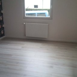 MKiP - Jasne panele podłogowe w pokoju po remoncie. Ściana z tapetą w kwiaty, okno z grzejnikiem. Widoczne listwy przypodłogowe i gładkie ściany.