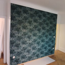 MKiP - Pokój po remoncie: ściana z tapetą w roślinny wzór, drewniana podłoga, białe drzwi. Widoczna drabina i narzędzia, co sugeruje prace wykończeniowe.