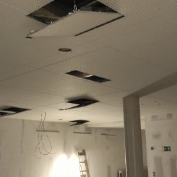 MKiP - Sufit podwieszany z odkrytymi fragmentami, widoczne kable i drabina. W tle ściany w trakcie szpachlowania. Prace wykończeniowe w pomieszczeniu.