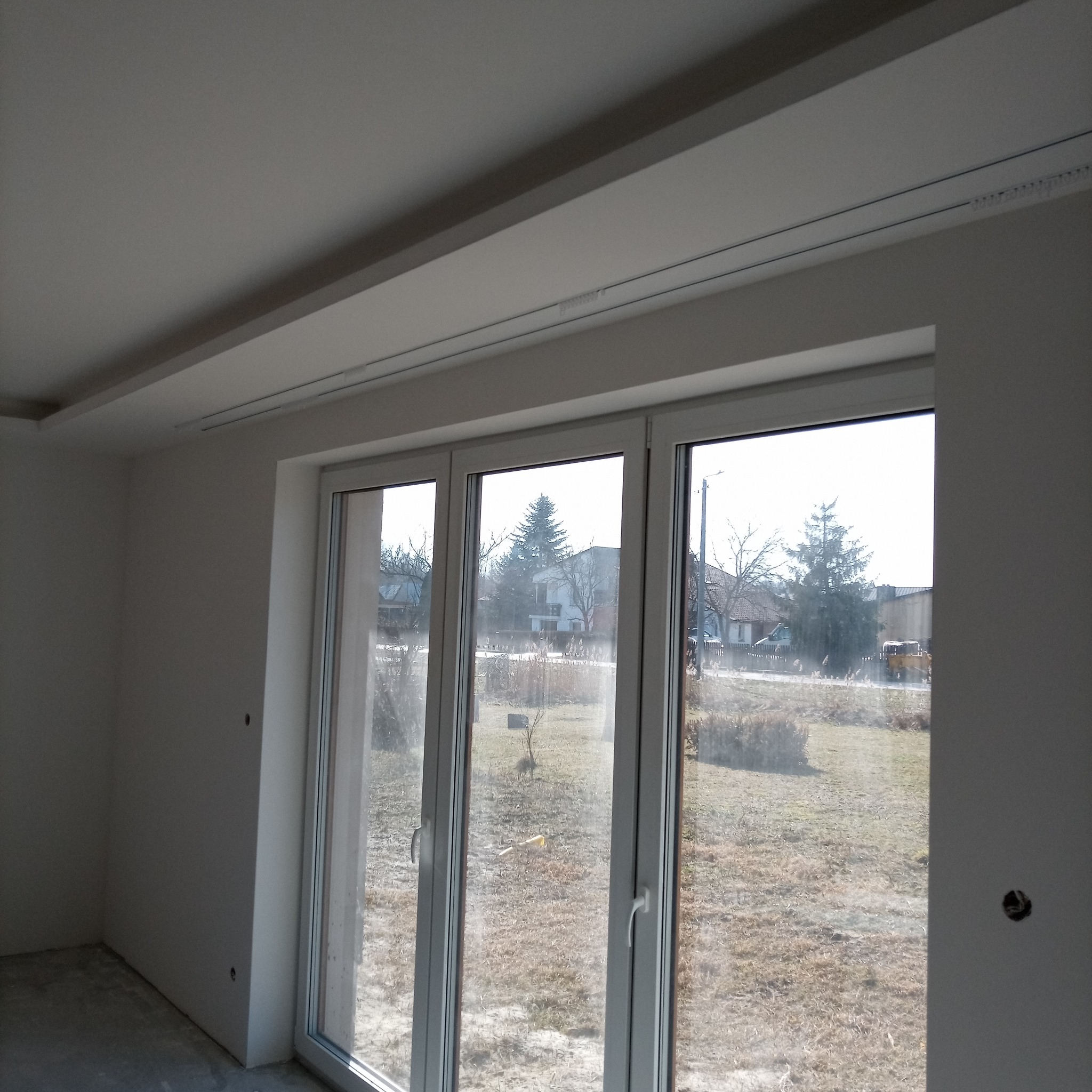 Jasne wnętrze po remoncie z dużym oknem balkonowym i karniszem sufitowym. Ściany i sufit pomalowane na biało, widoczne ślady po gładzi.