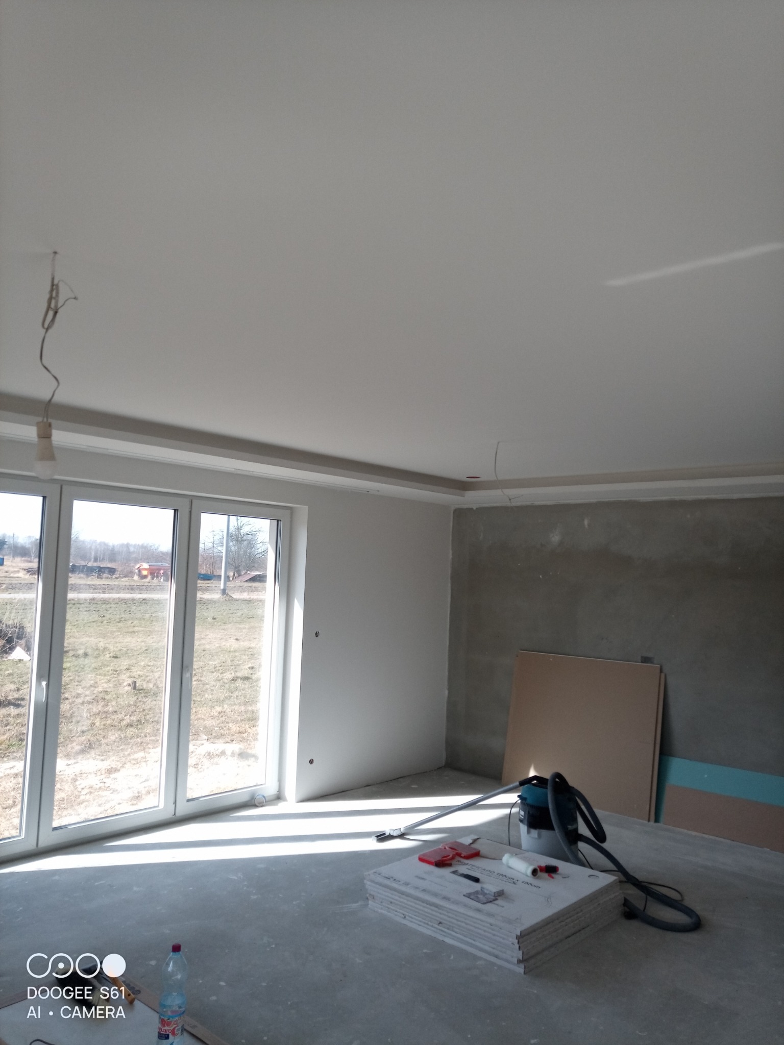 Wnętrze w trakcie prac wykończeniowych: surowa ściana, płyty gipsowe, narzędzia, okno z widokiem na zewnątrz. Widoczne przygotowania do dalszych prac.