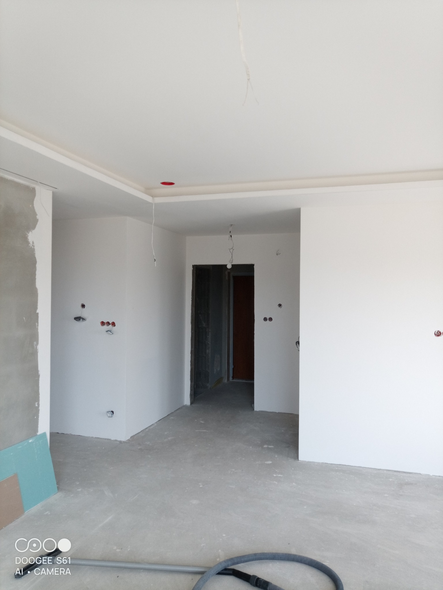 Wnętrze w trakcie wykańczania: białe ściany z widocznymi puszkami elektrycznymi, surowy beton na podłodze, płyta gipsowa i odkurzacz przemysłowy w rogu.