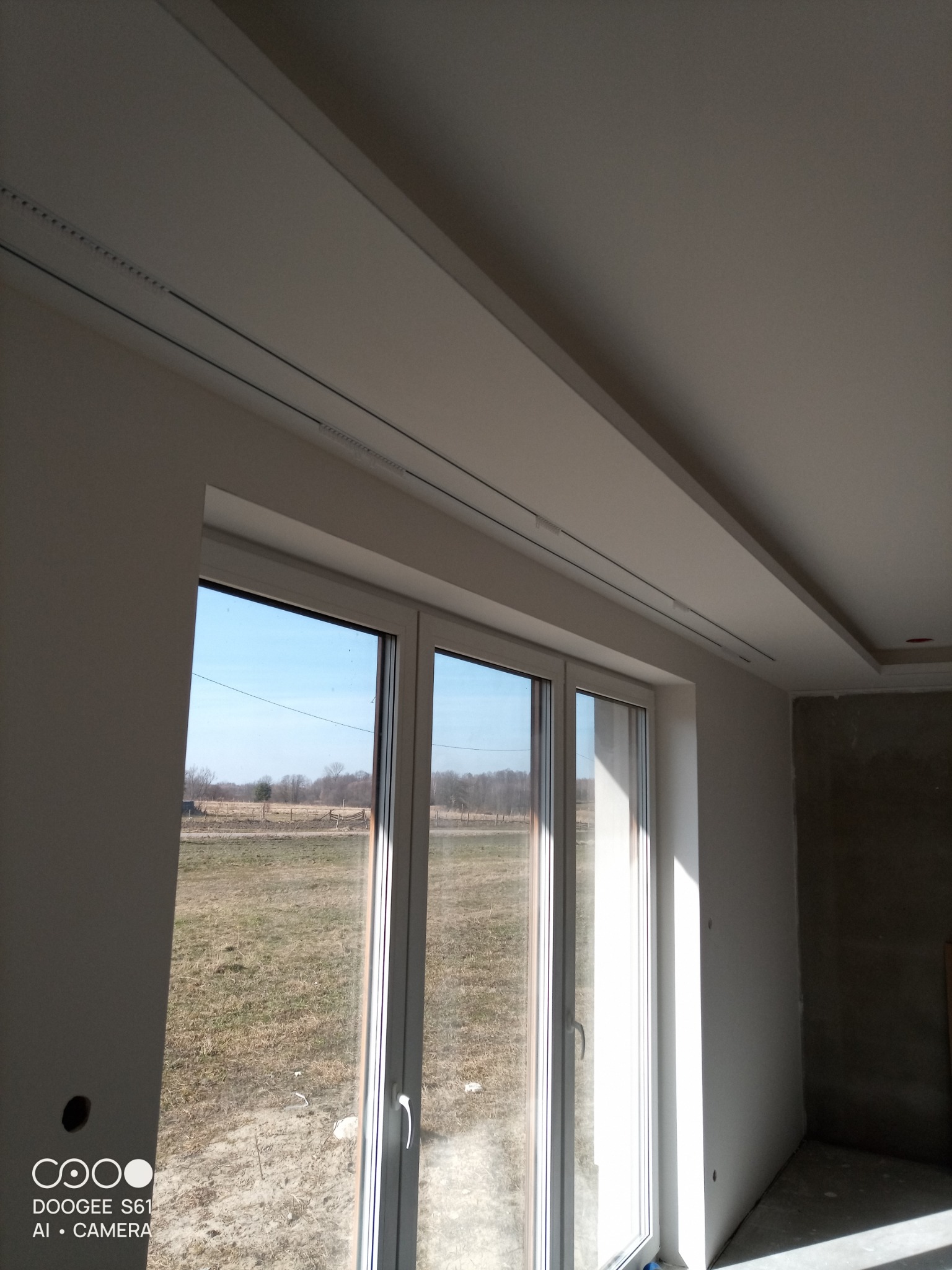 Białe, wykończone wnętrze z oknem i podwieszanym sufitem z oświetleniem szynowym. Ściany przygotowane do malowania, widok na zewnątrz przez okno.